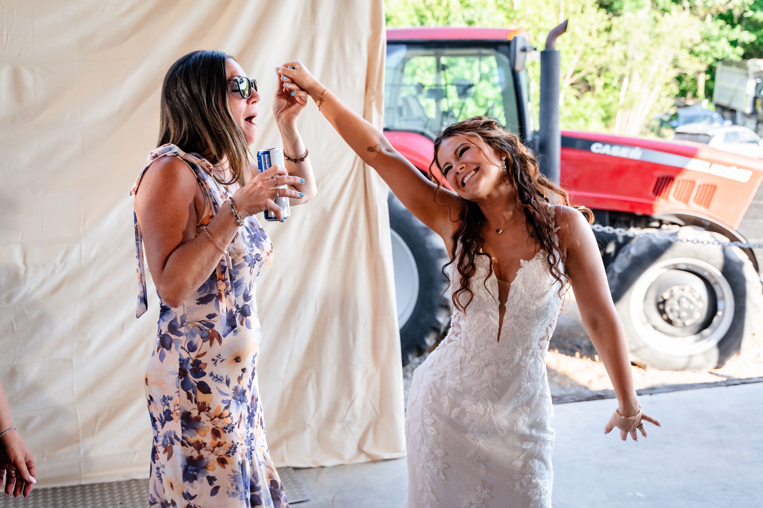 Bride dancing joyfully with a guest in a floral dress at an outdoor farm wedding celebration.