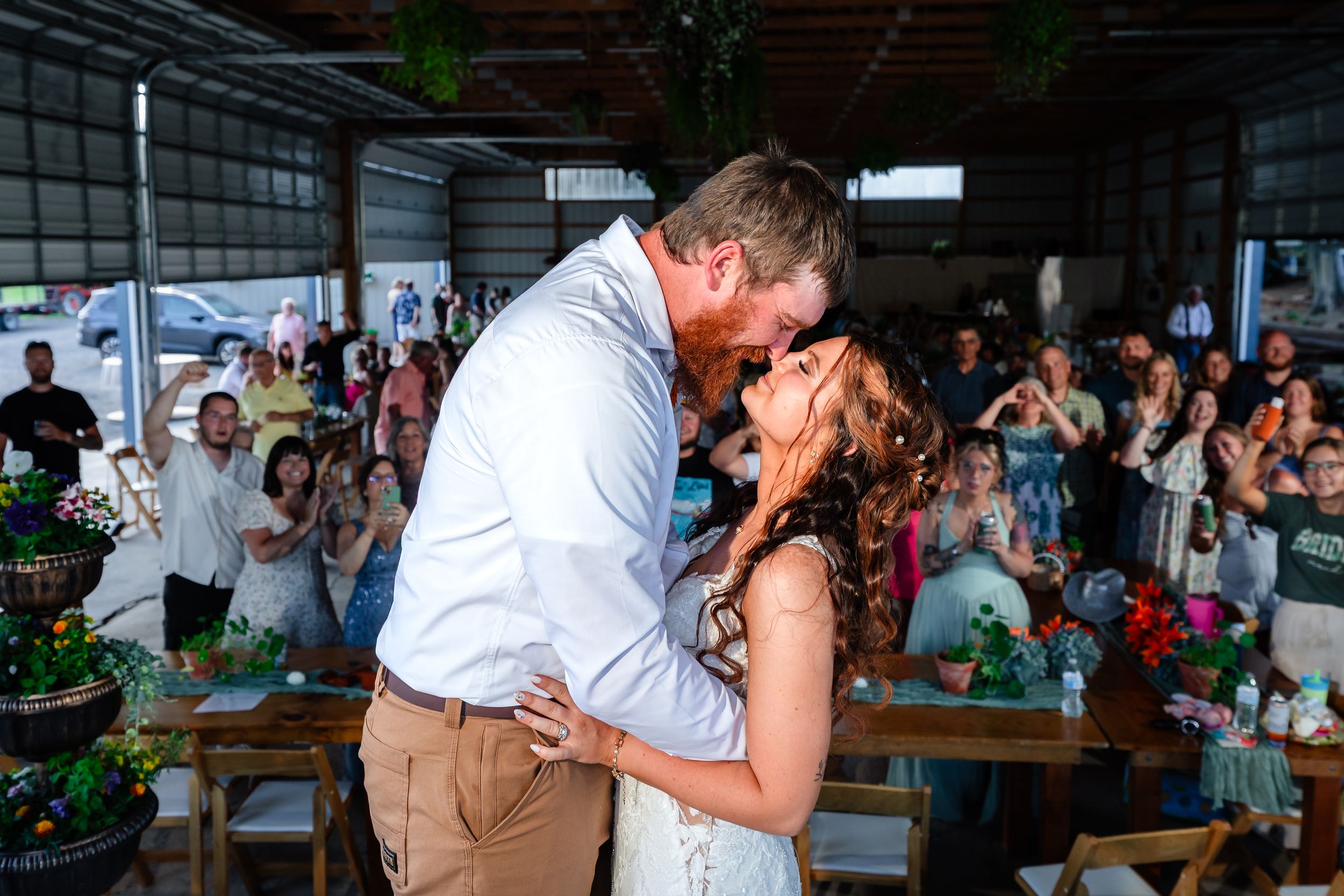 Bride and groom share their first kiss at a rustic barn wedding reception while guests cheer and applaud.