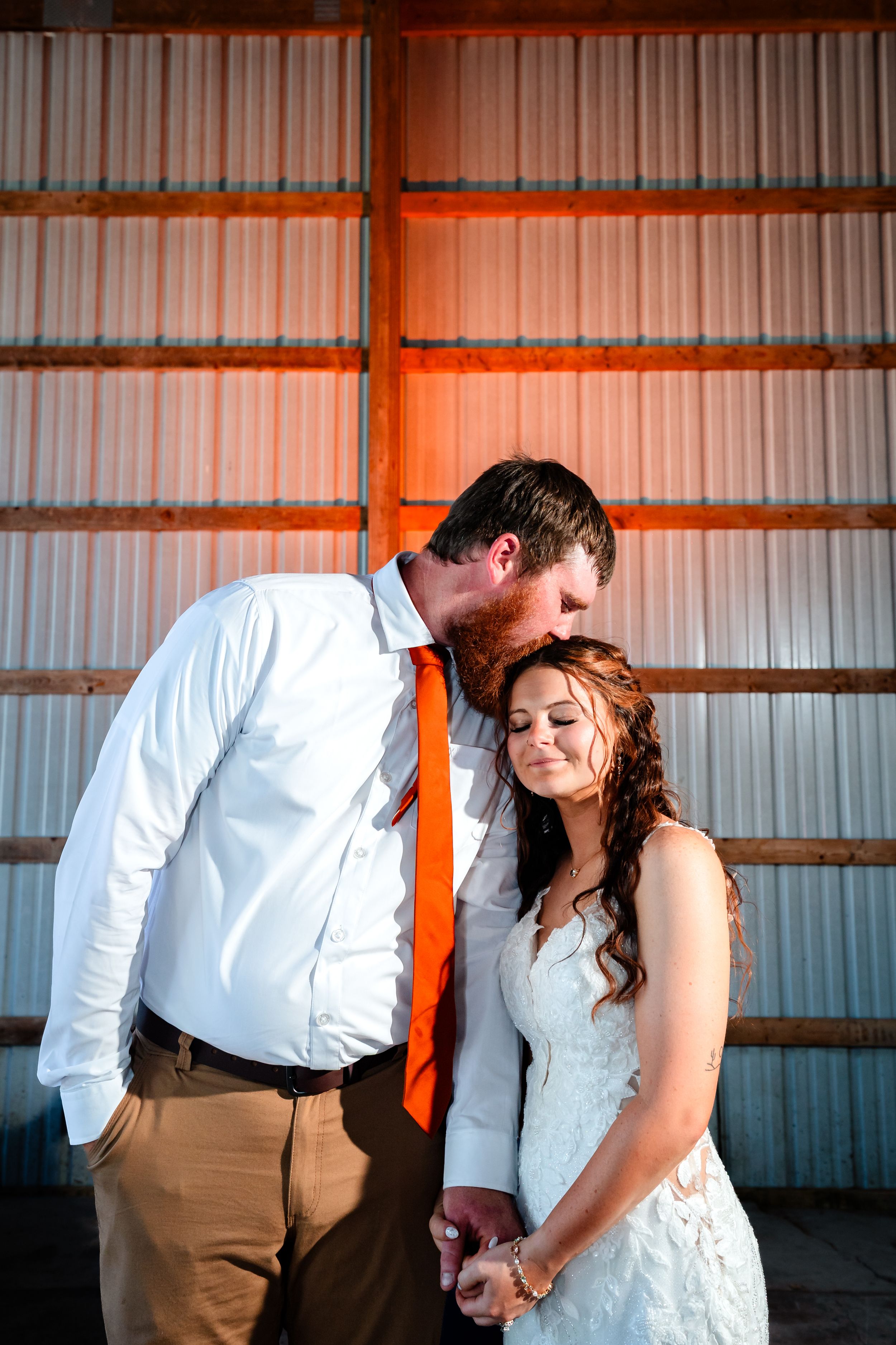 Bride and groom share a tender moment in a barn, groom kissing her head while holding hands in golden light.