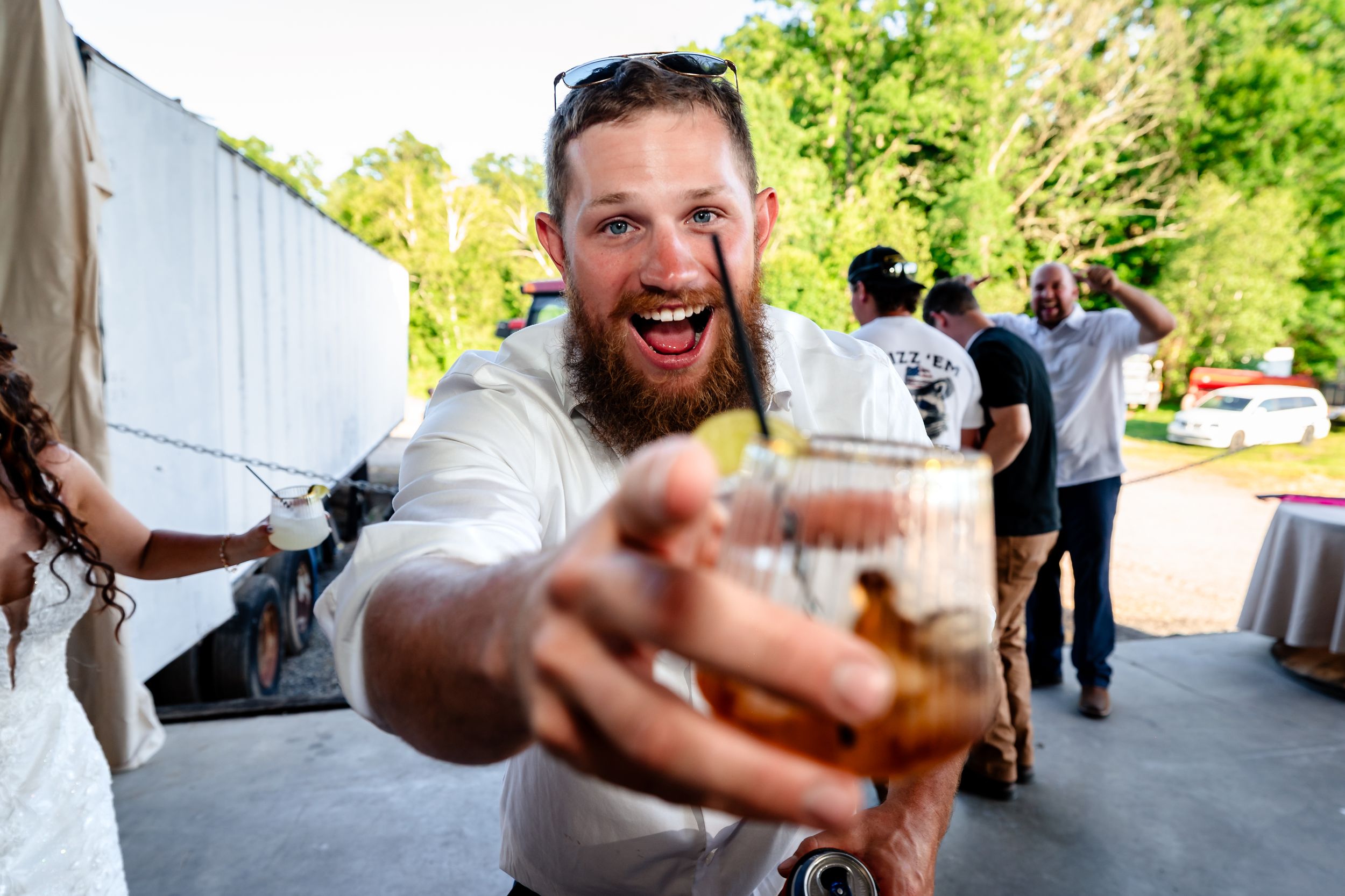 Excited bearded man in white shirt holds up a drink toward the camera at an outdoor wedding reception.