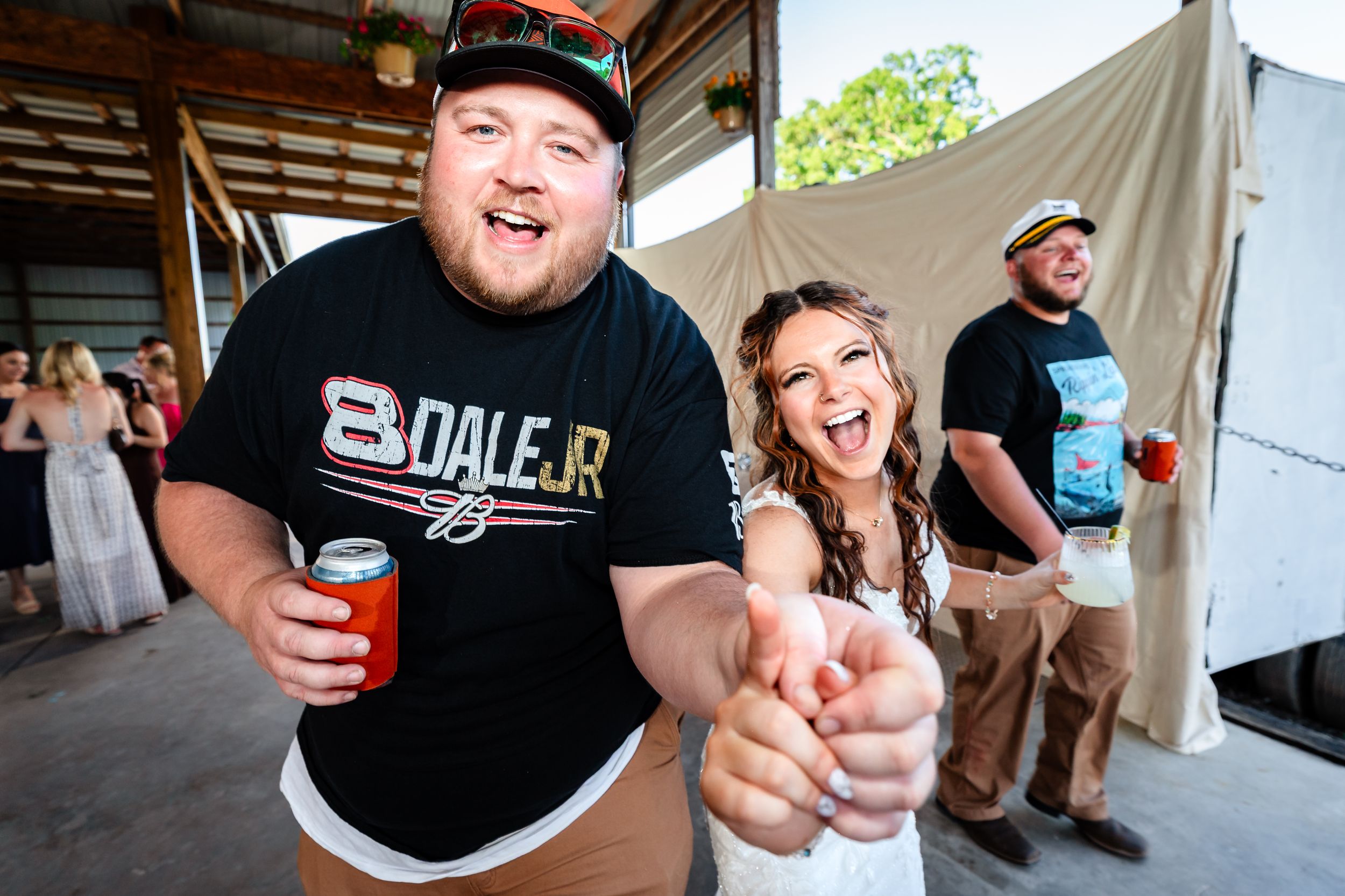 Man in Dale Jr shirt and woman laughing pointing at camera at rustic outdoor wedding reception.
