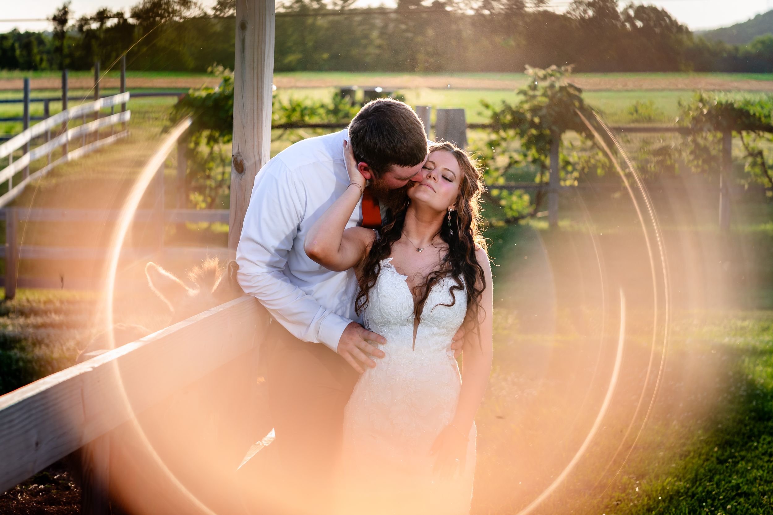Couple sharing a romantic kiss at golden hour on a farm wedding venue with lens flare.