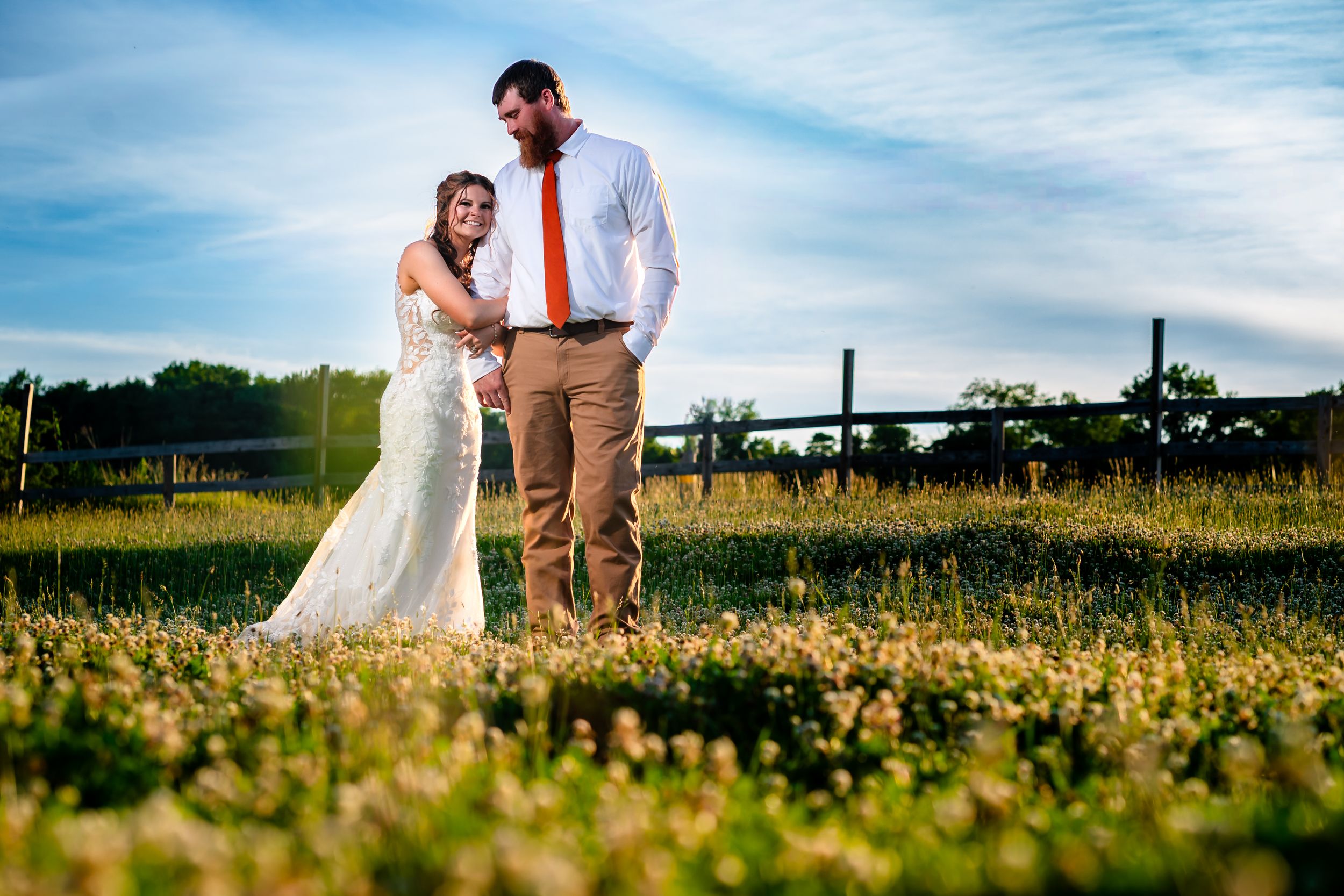 Bride and groom pose romantically in a golden sunlit field with a wooden fence backdrop at sunset.