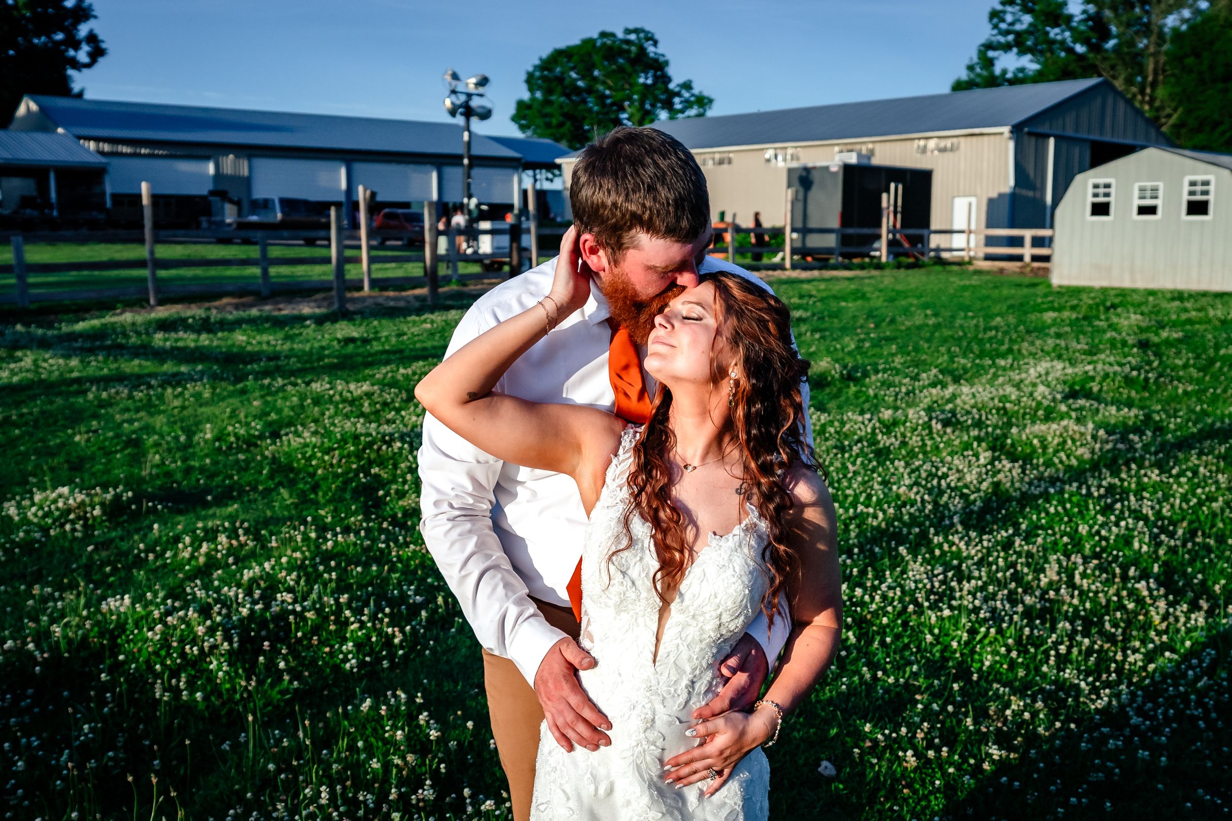 Bride and groom embracing in green field at golden hour near farm buildings at rustic wedding.