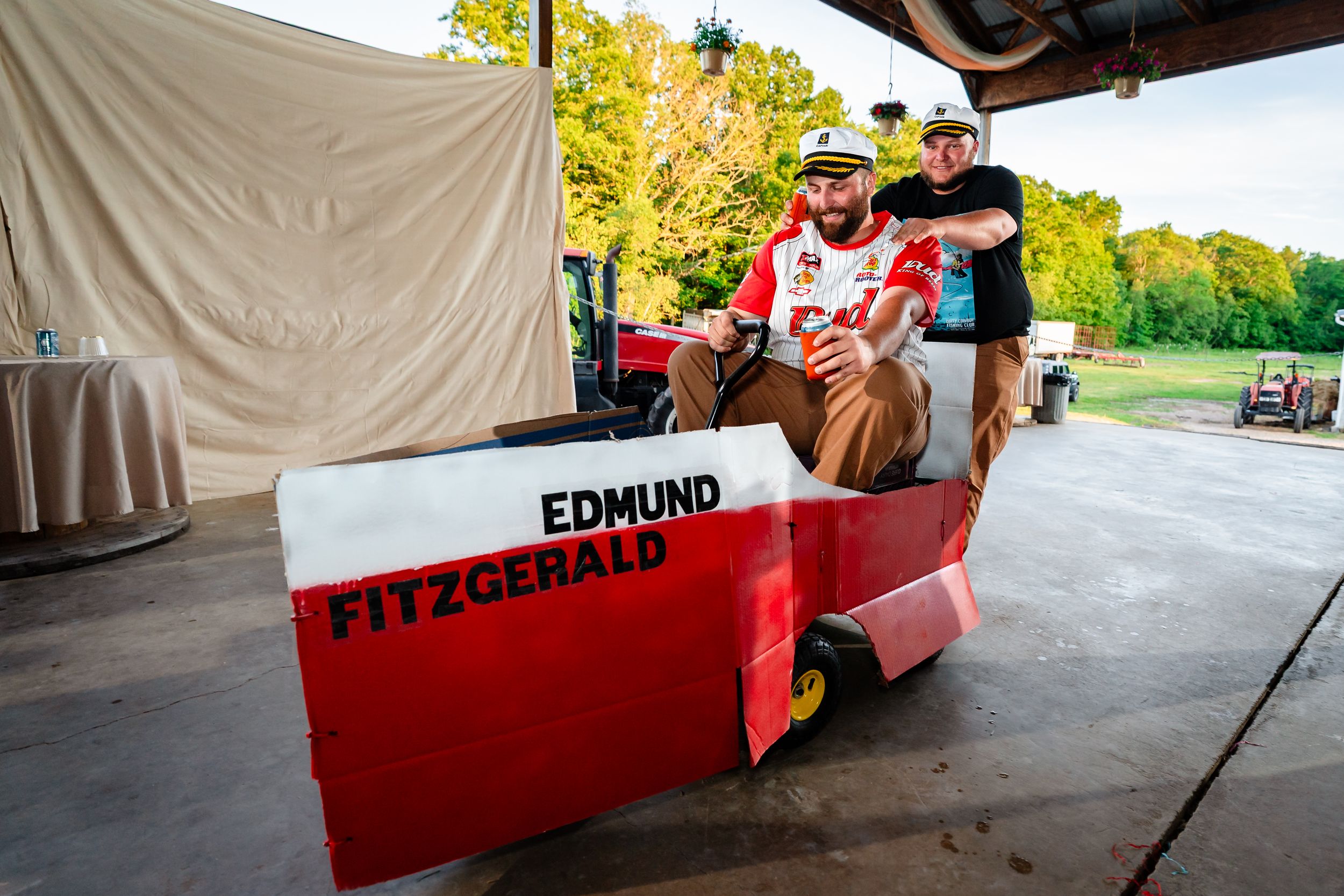 Two men pose with a red and white Edmund Fitzgerald themed go-kart at an outdoor event.