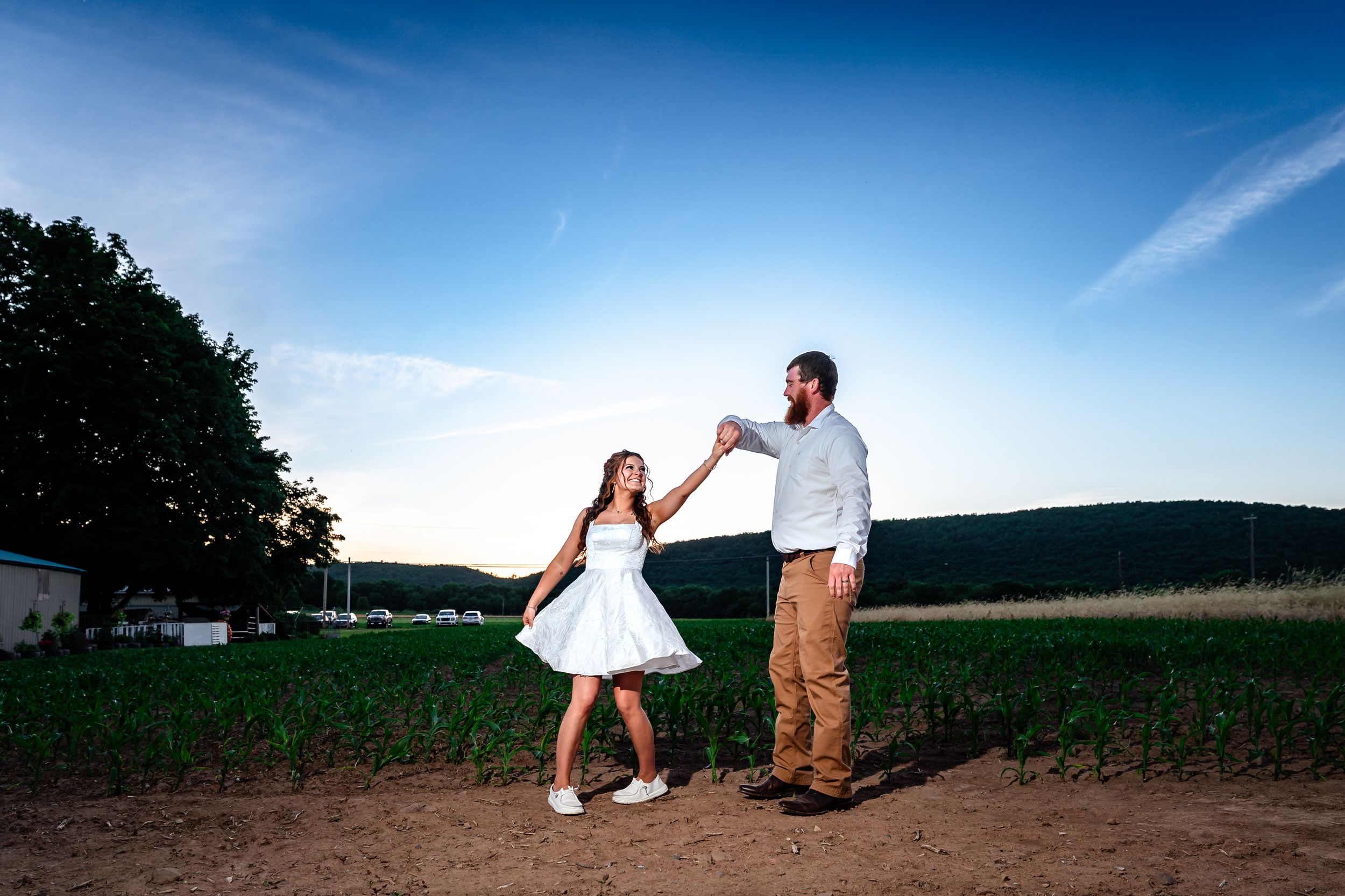 Couple dancing joyfully in a field at dusk with lush green crops and trees in the background.