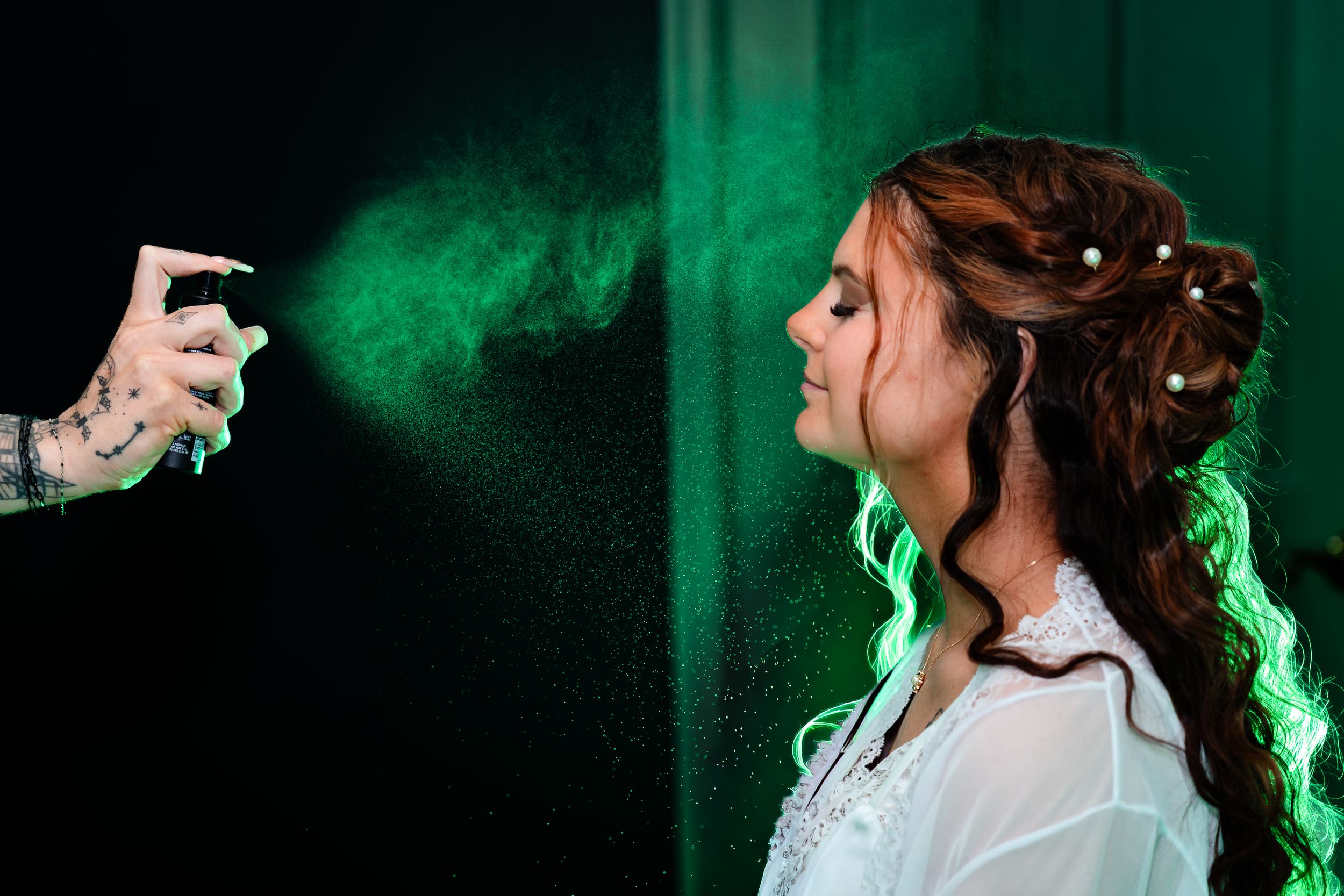 A bride has green color spray misted onto her face during wedding day preparations against a dark backdrop.