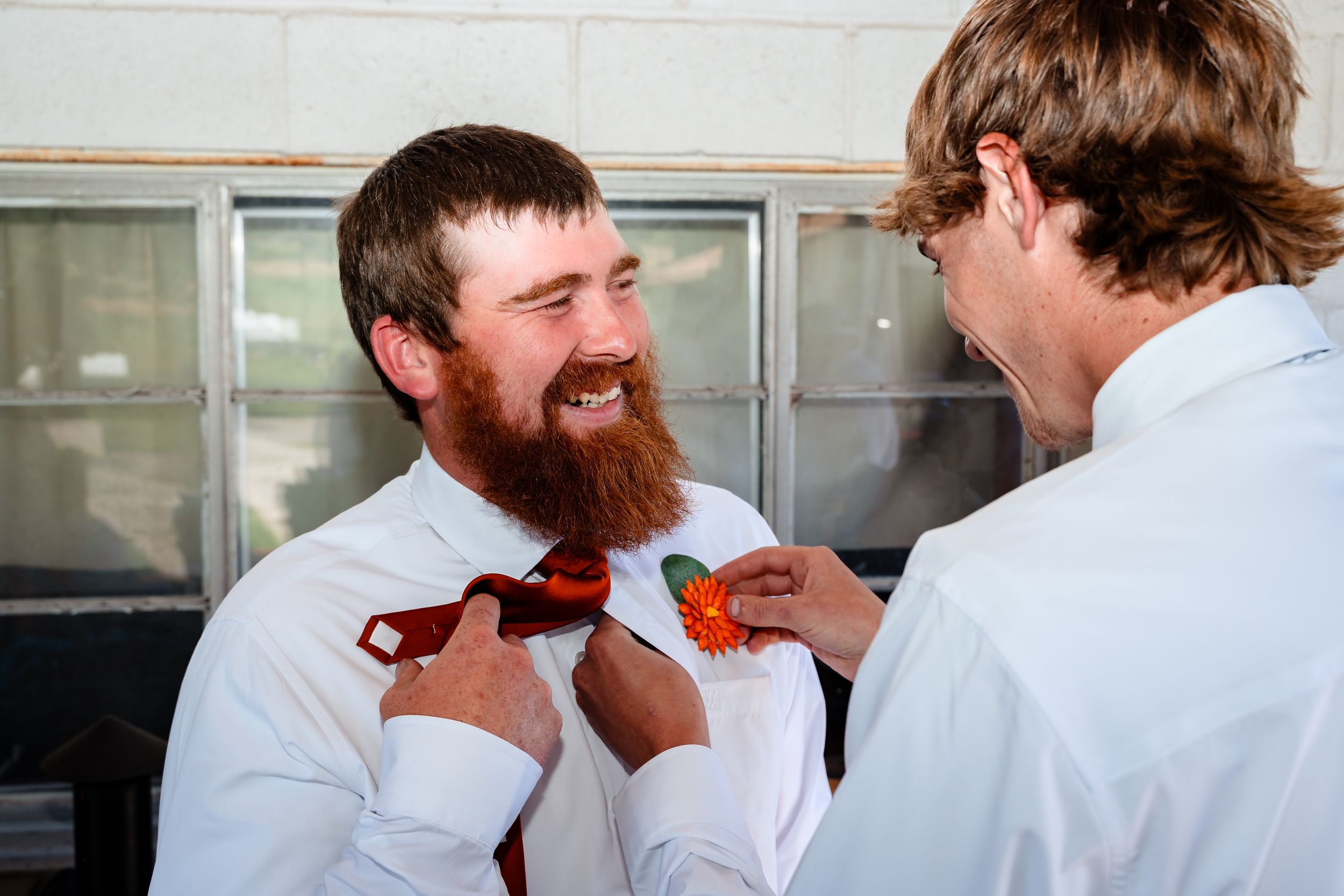 Groom with red beard laughs as groomsman pins orange boutonniere on his red tie outdoors.