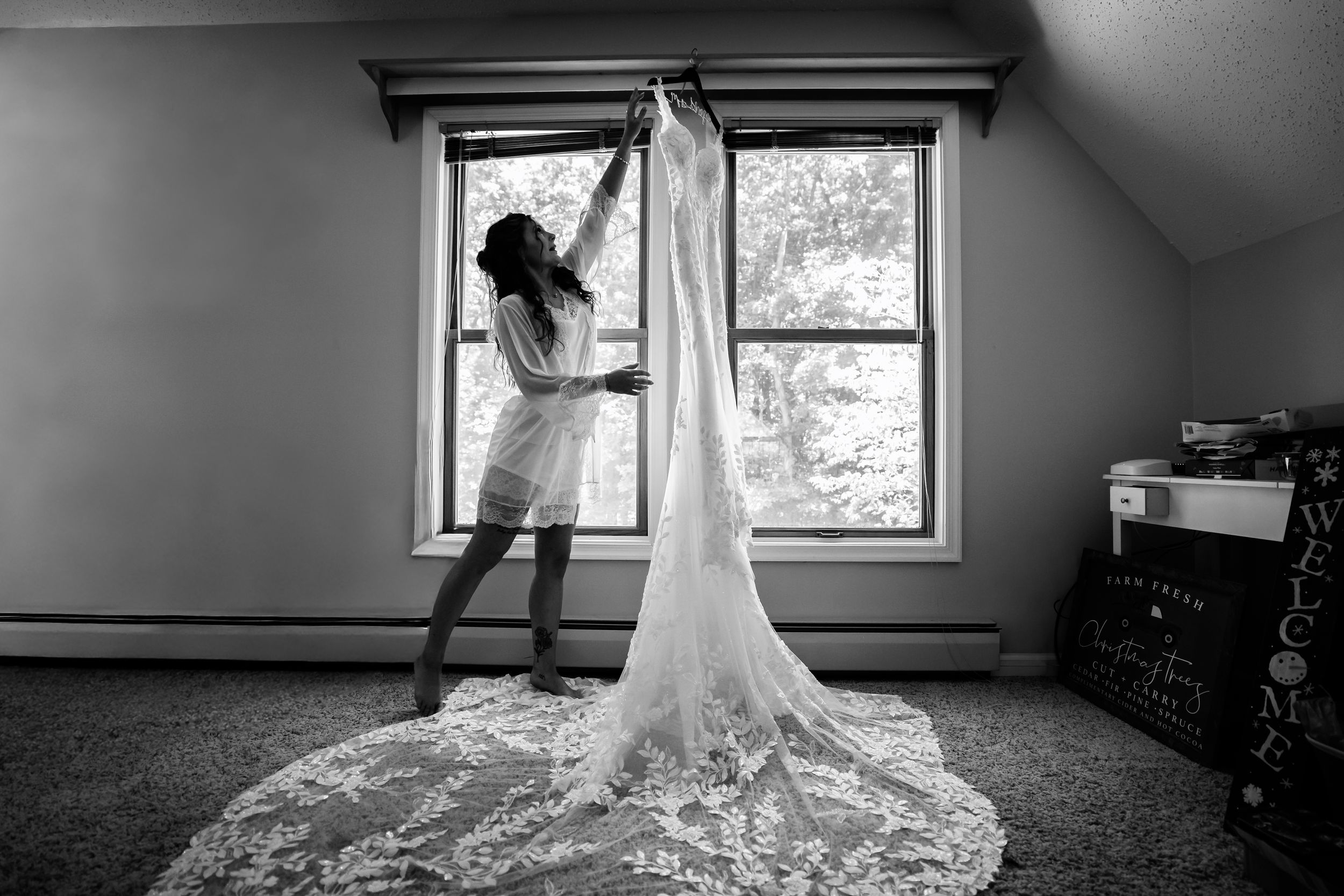 Black and white silhouette of bride holding up long lace wedding gown in front of bright window.