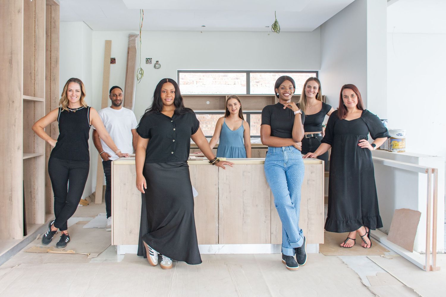 Seven interior design professionals in black outfits pose together in a modern, minimalist kitchen space under renovation.