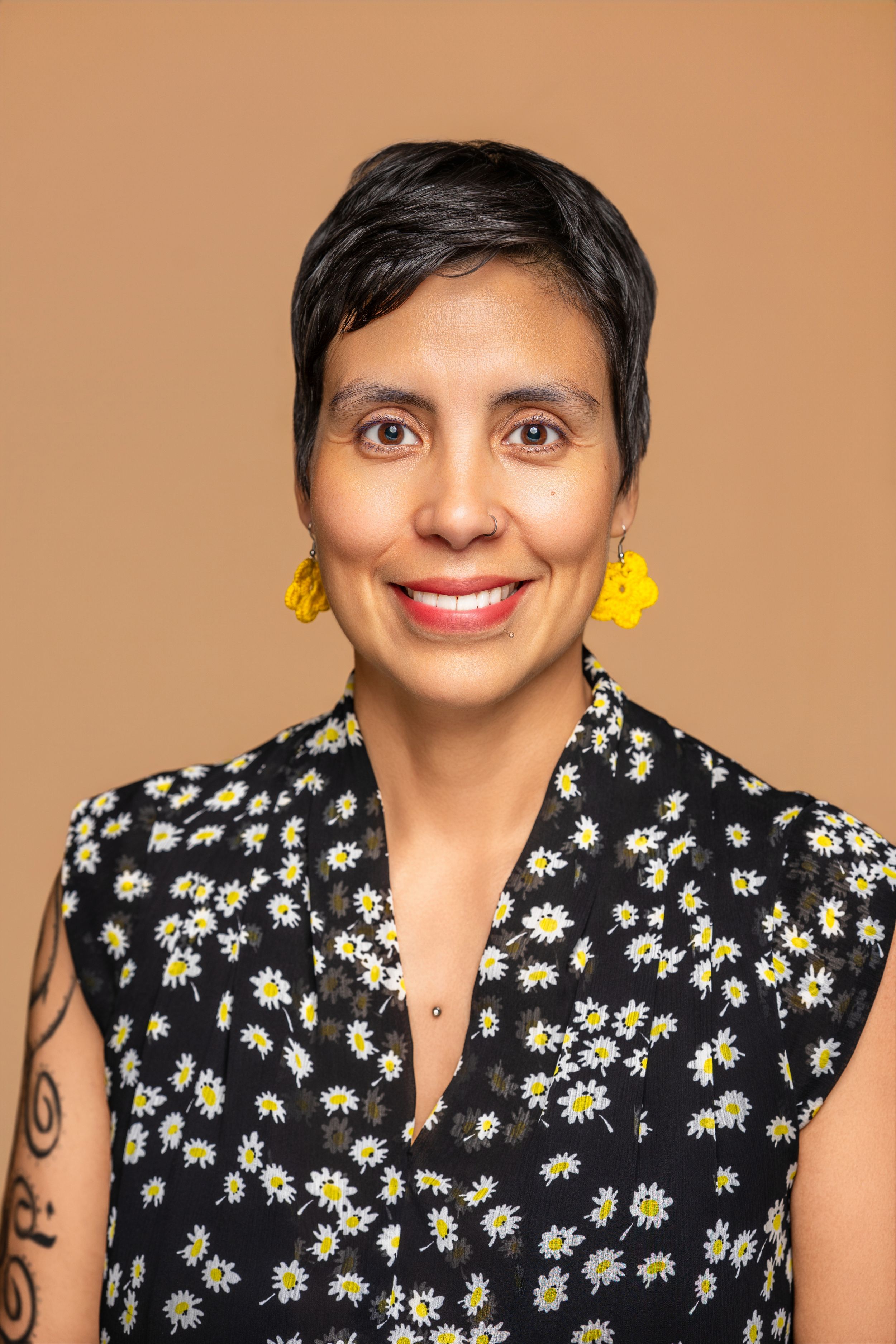 Smiling woman with short dark hair wearing yellow earrings and floral dress outdoors in forest setting.