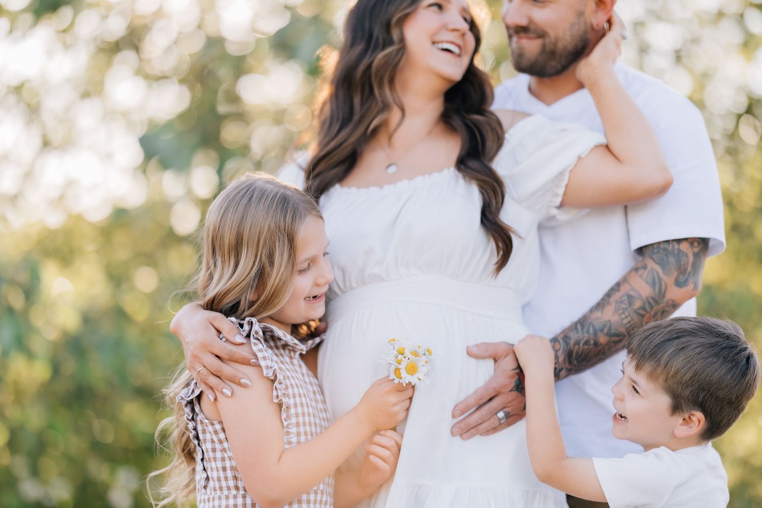 Pregnant woman in white dress with tattooed husband and two children touching her baby bump outdoors.