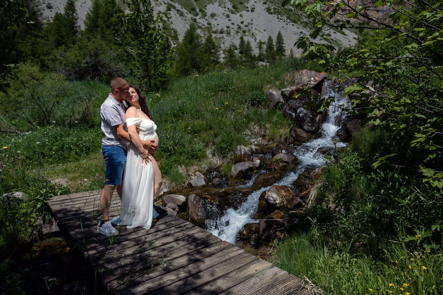 Couple embracing on wooden bridge by mountain waterfall surrounded by lush green trees during outdoor maternity photoshoot.
