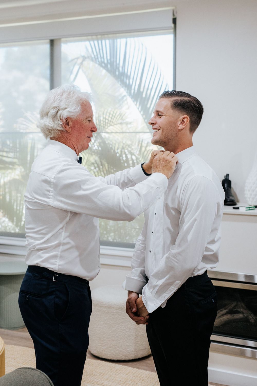Elderly father helping young groom button his white dress shirt while getting ready for wedding ceremony.