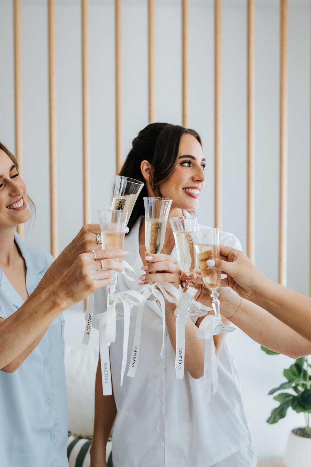 Women in white robes toasting with champagne glasses, smiling and celebrating at a bridal party gathering.