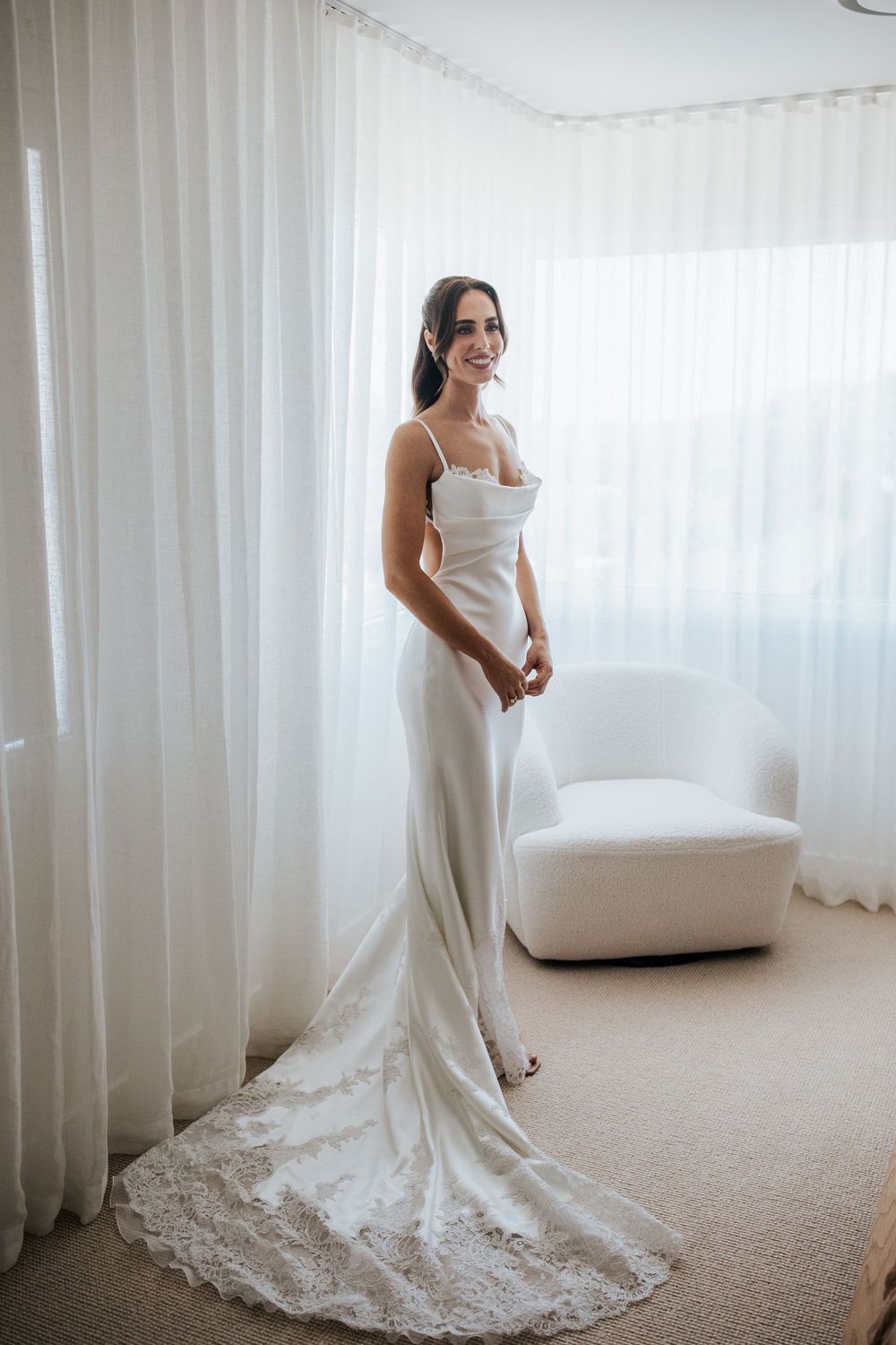 Bride in elegant white satin slip wedding gown with lace train, smiling by sheer curtains in bright room.