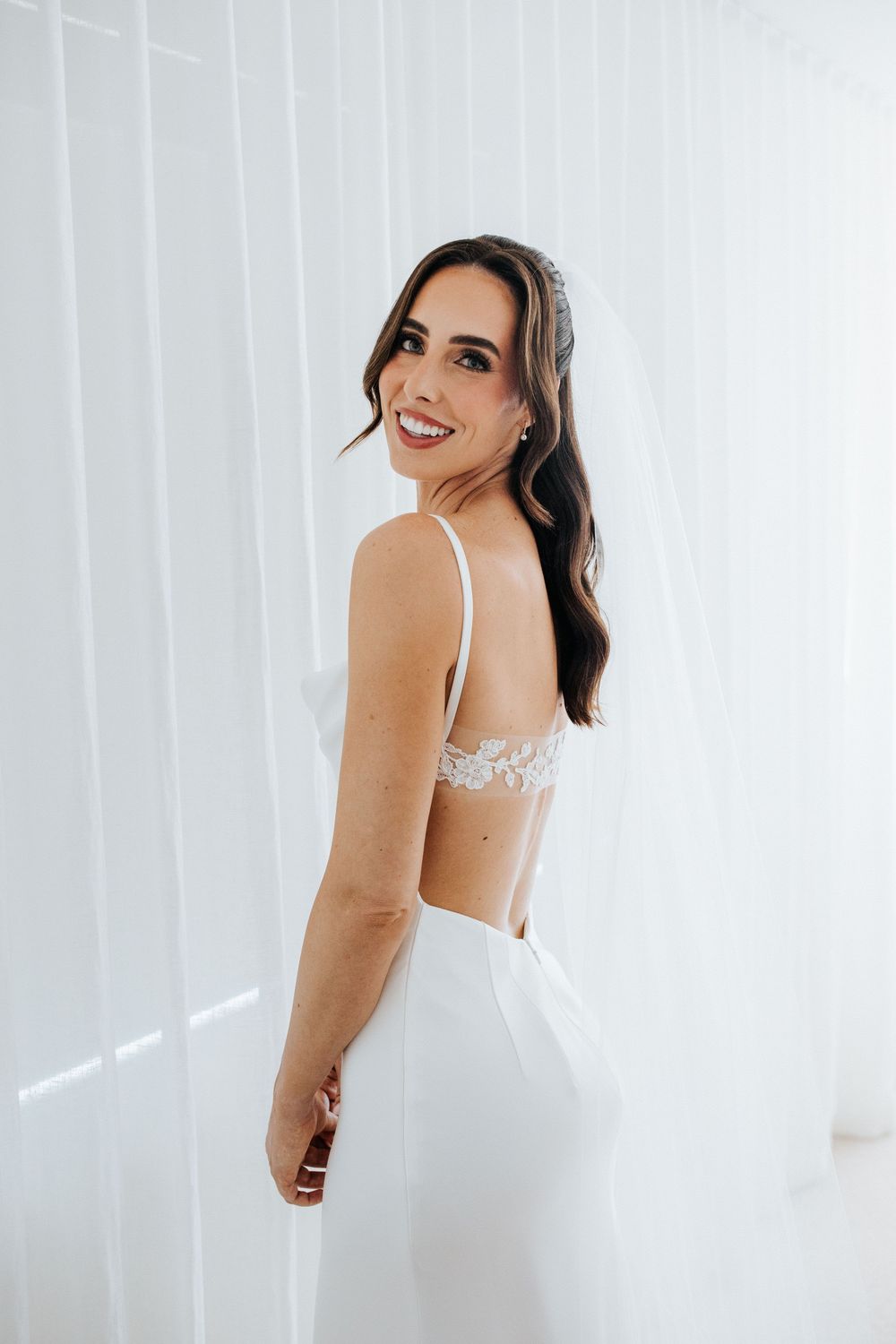 Smiling bride in backless white wedding gown with lace detail and veil, posing elegantly against white backdrop.