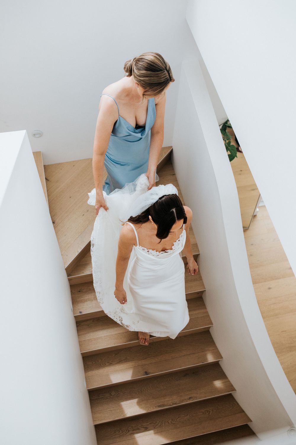 Bride in white gown walks down wooden stairs assisted by bridesmaid in blue satin dress.