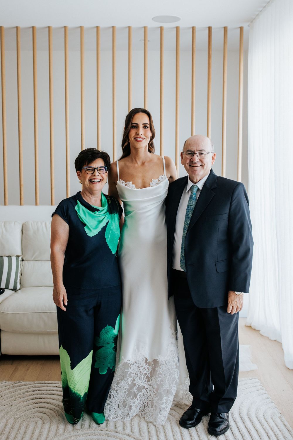 Bride in white lace gown poses with parents in elegant room, mother in green floral outfit, father in navy suit.