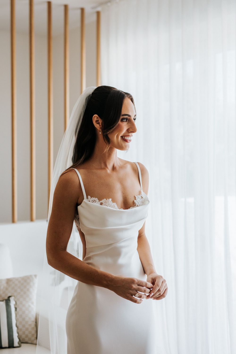 Smiling bride in a white satin cowl neck gown with lace detail and veil, standing by a bright window.