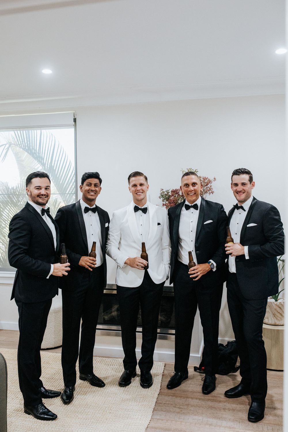 Five groomsmen in black tuxedos and bow ties, with groom in white jacket, holding beer bottles at wedding.