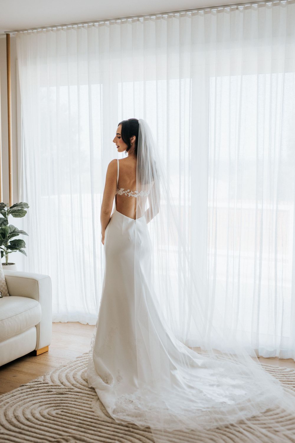 Bride in elegant backless white gown with veil stands before sheer curtained window in bright modern room.