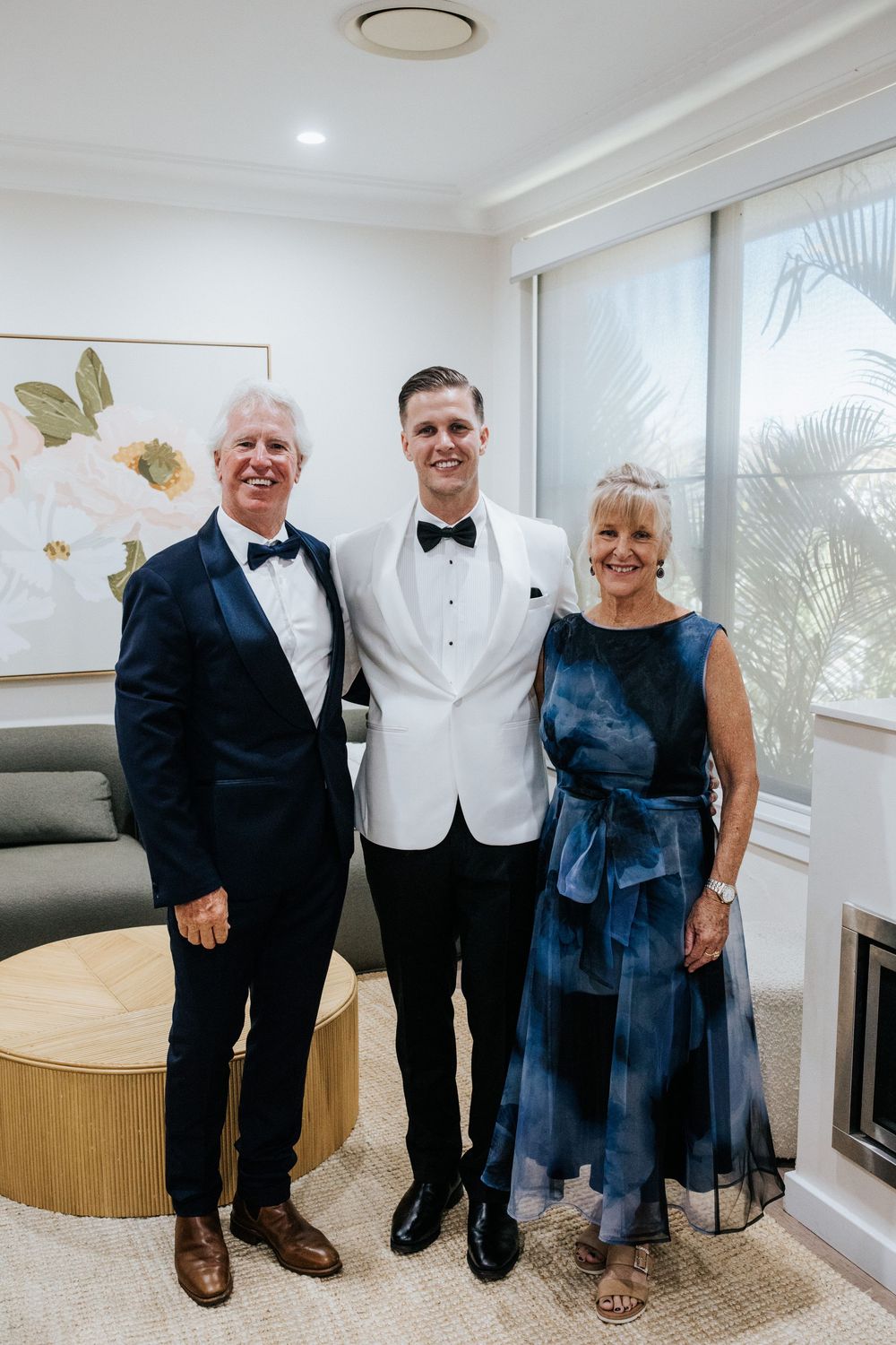 A groom in a white tuxedo poses with his parents in formal attire in an elegant room before a wedding.