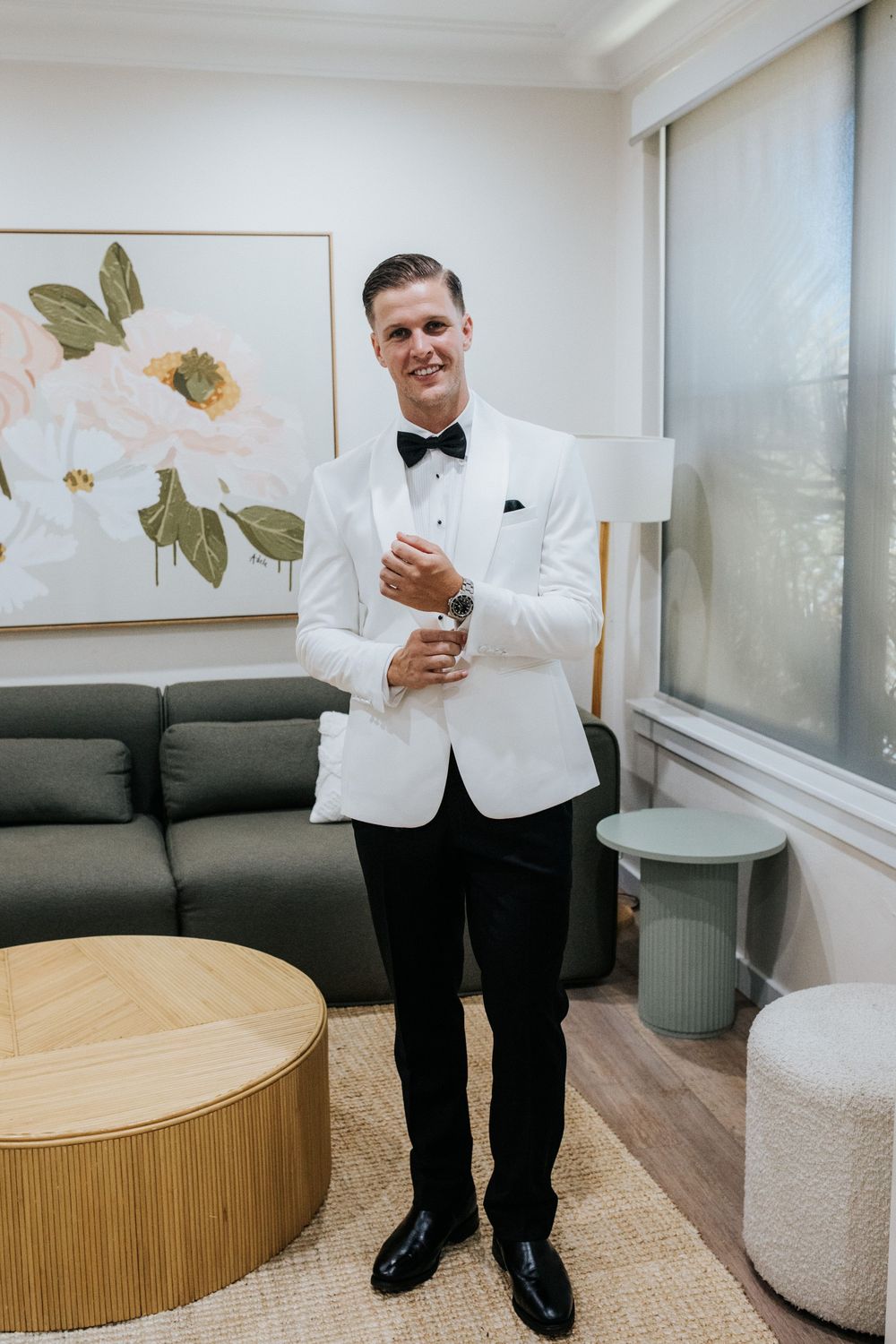 Groom in white tuxedo jacket and black bow tie adjusting cufflinks in elegant room with floral artwork.