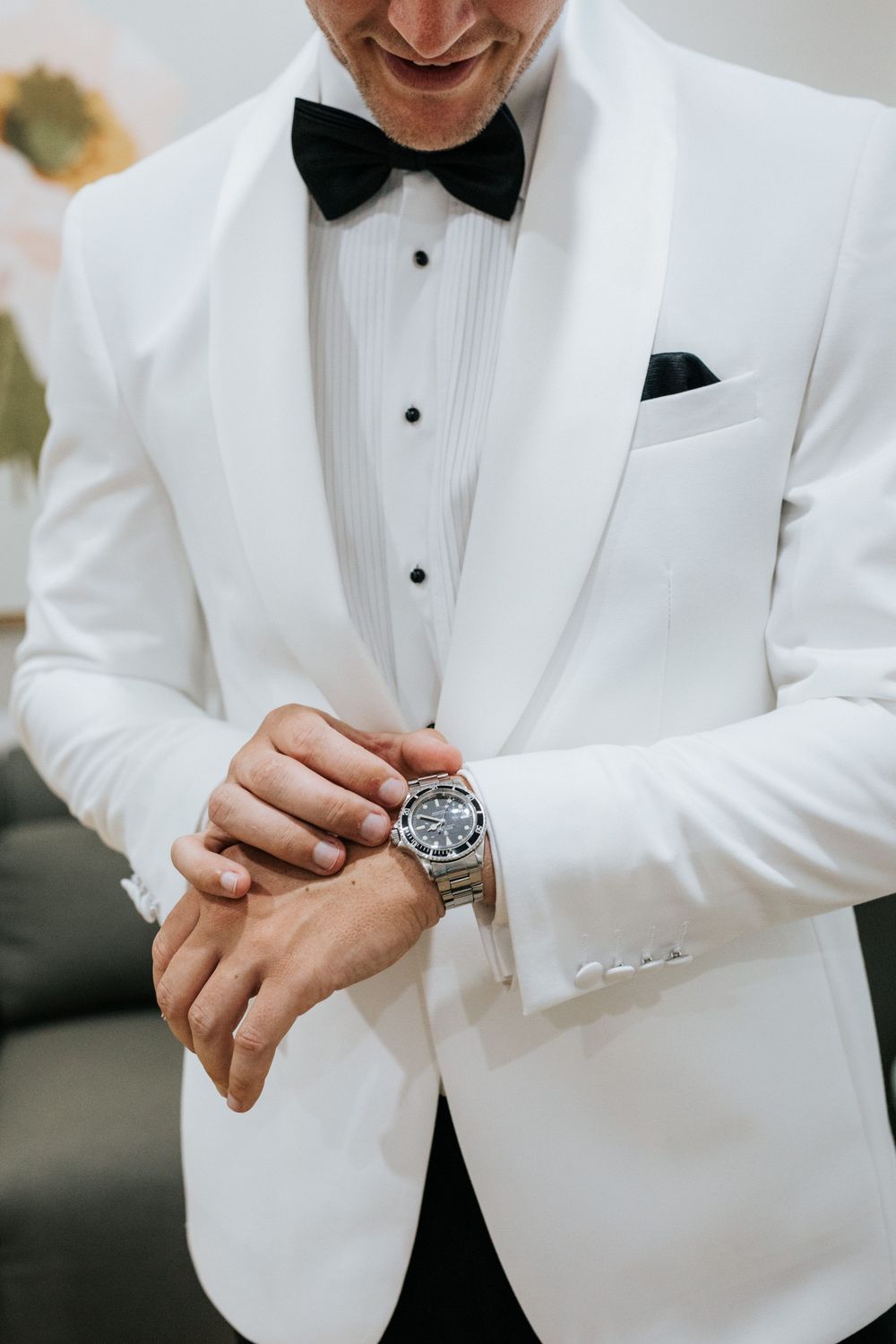 Smiling groom in white tuxedo jacket with black bow tie adjusting his silver watch before wedding ceremony.