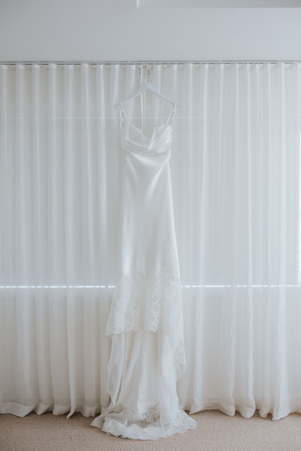 Elegant white satin wedding dress with lace hem hanging on a hanger against sheer white curtains.