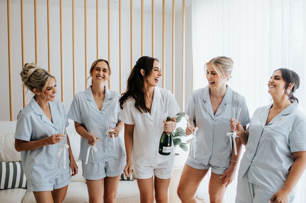 Five bridesmaids in matching light blue pajamas celebrate with champagne, laughing and smiling together before a wedding.