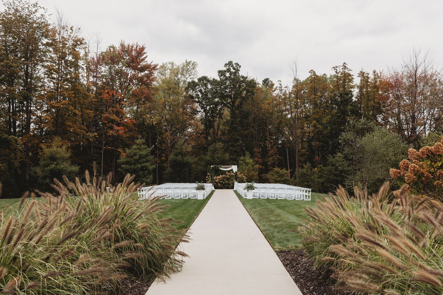 Elegant outdoor wedding ceremony setup with white chairs lining a path, surrounded by autumn trees in vibrant fall colors.