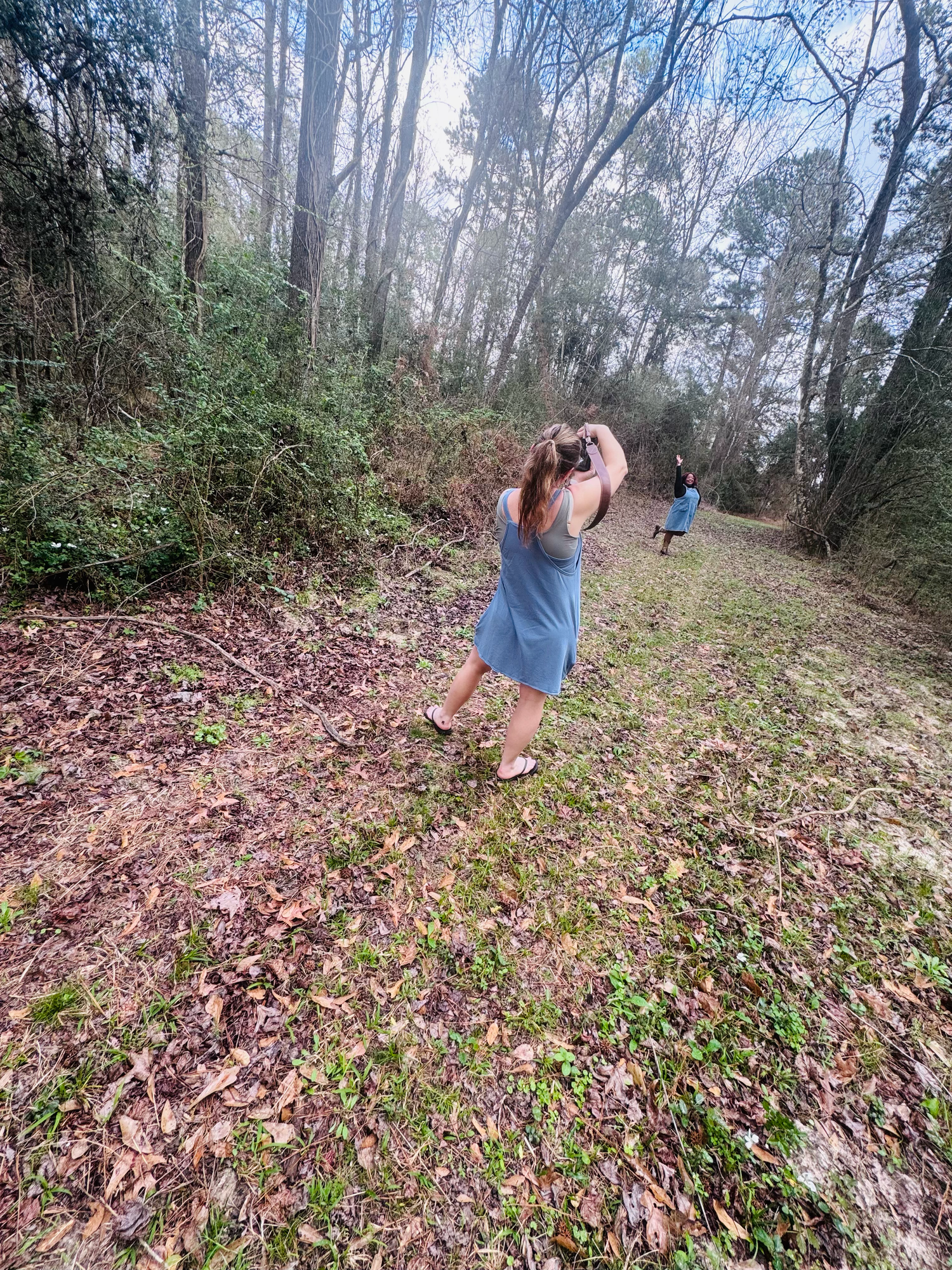 Two women walking on a wooded trail path surrounded by tall trees and fallen leaves in a misty forest.