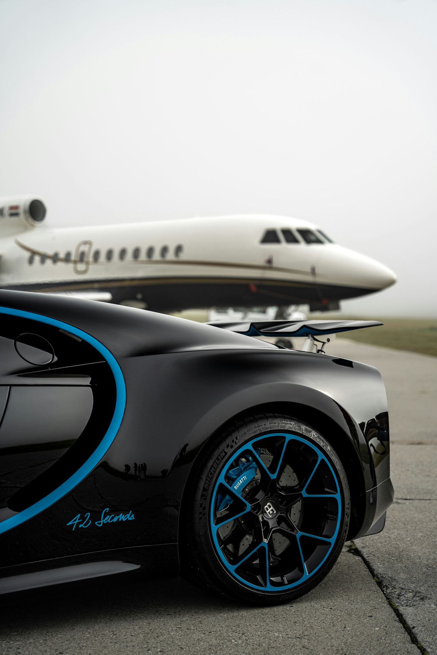 Black and blue Bugatti Chiron supercar on tarmac with a private jet in the misty background.