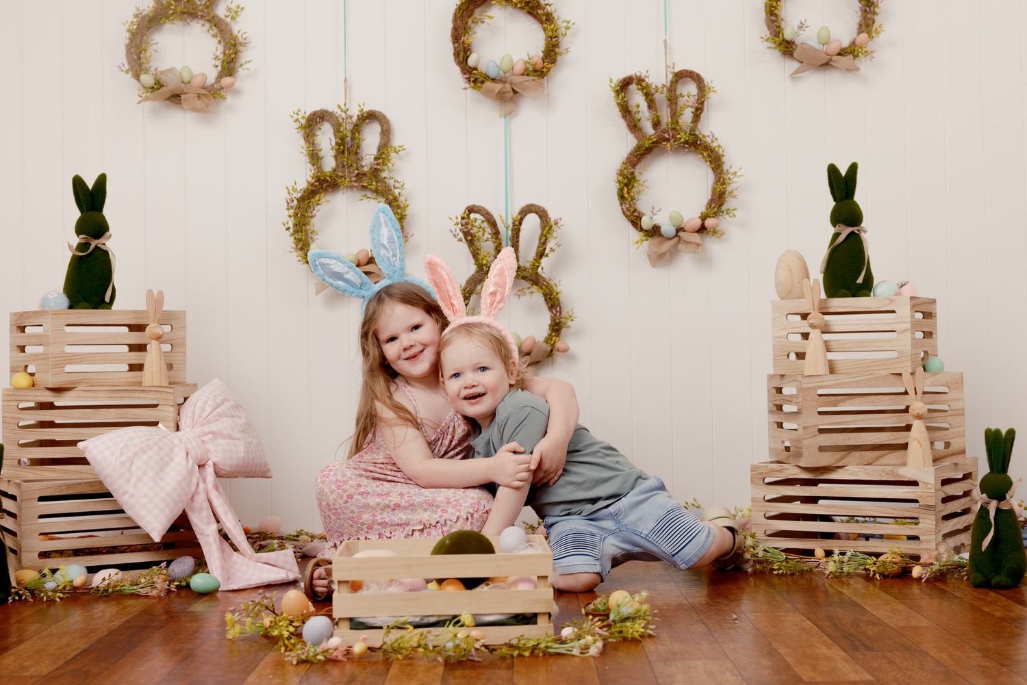 Two children wearing bunny ears pose among wooden crates and Easter decorations with wreaths and bunny figures on a white wall.