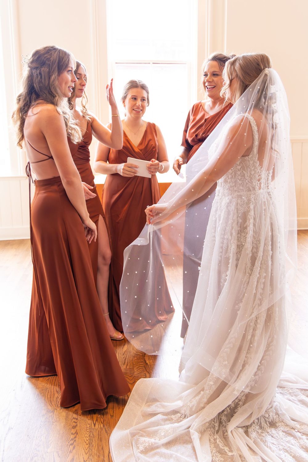 Bride in white lace gown and veil shares a joyful moment with bridesmaids in rust-colored dresses.