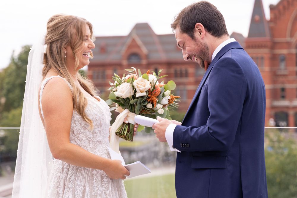 Bride in lace gown holding floral bouquet exchanges vows with groom in navy suit outdoors at the Alcove Cincinnati wedding venue.