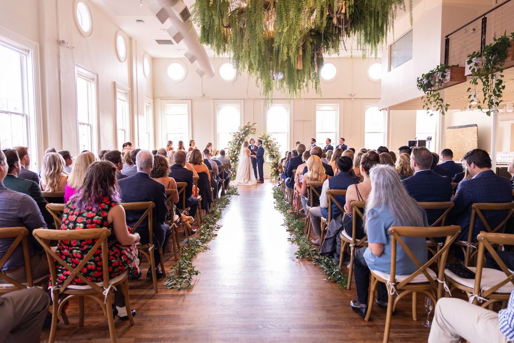 Elegant indoor wedding ceremony at the Alcove Cincinnati with guests seated along a floral aisle, lush greenery hanging from the ceiling.