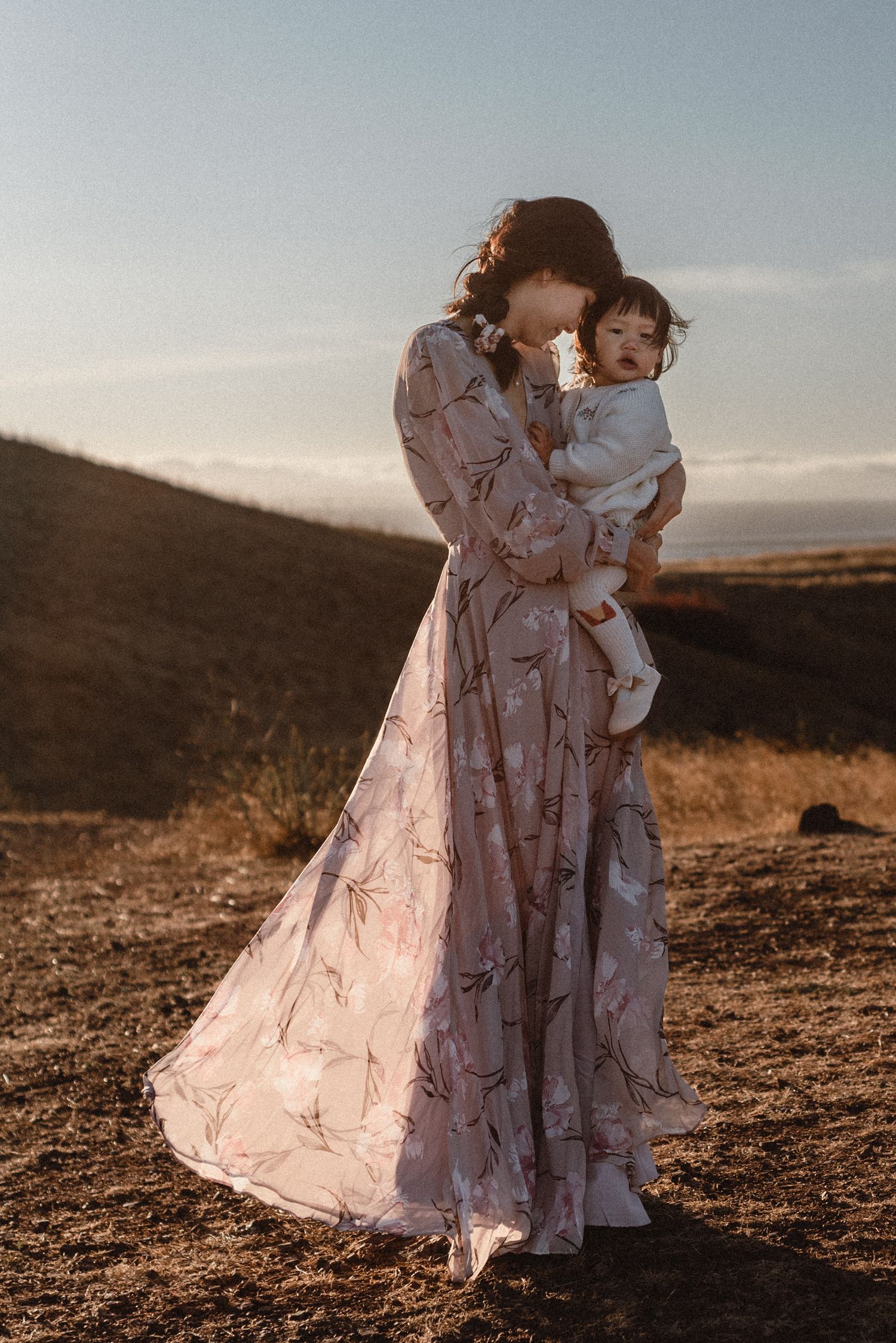 Mother in floral gown holds baby at golden hour in scenic outdoor landscape.