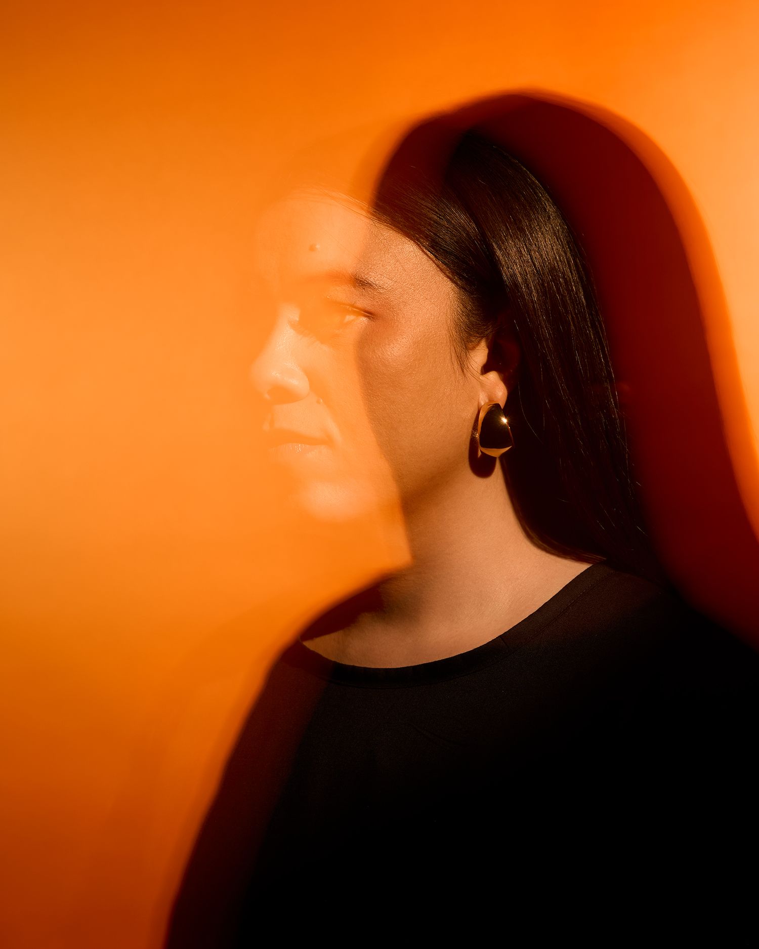 A woman with dark hair poses against an orange background with a dramatic double exposure shadow effect on her face.