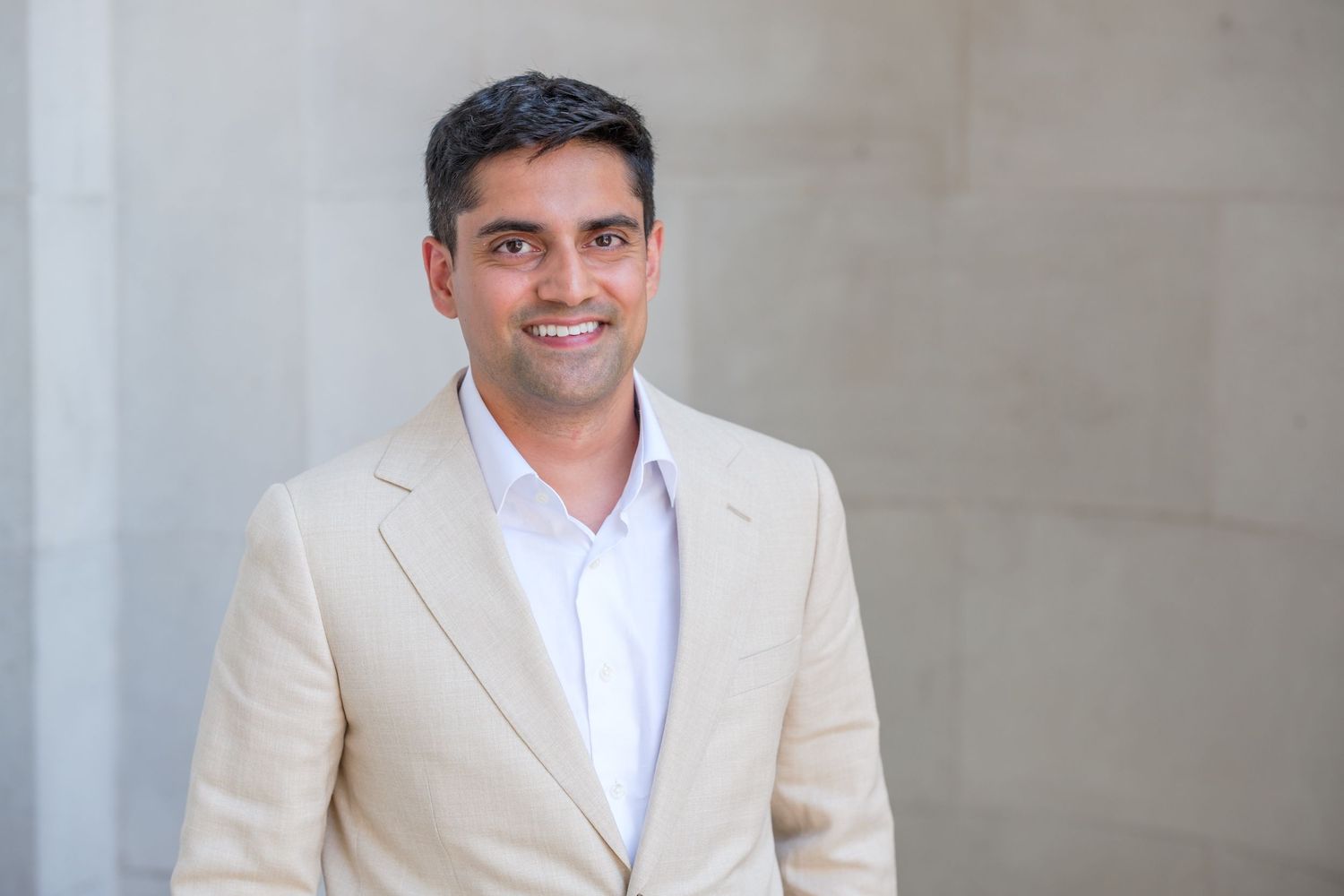 Professional man in beige blazer smiling confidently against a modern gray wall backdrop.