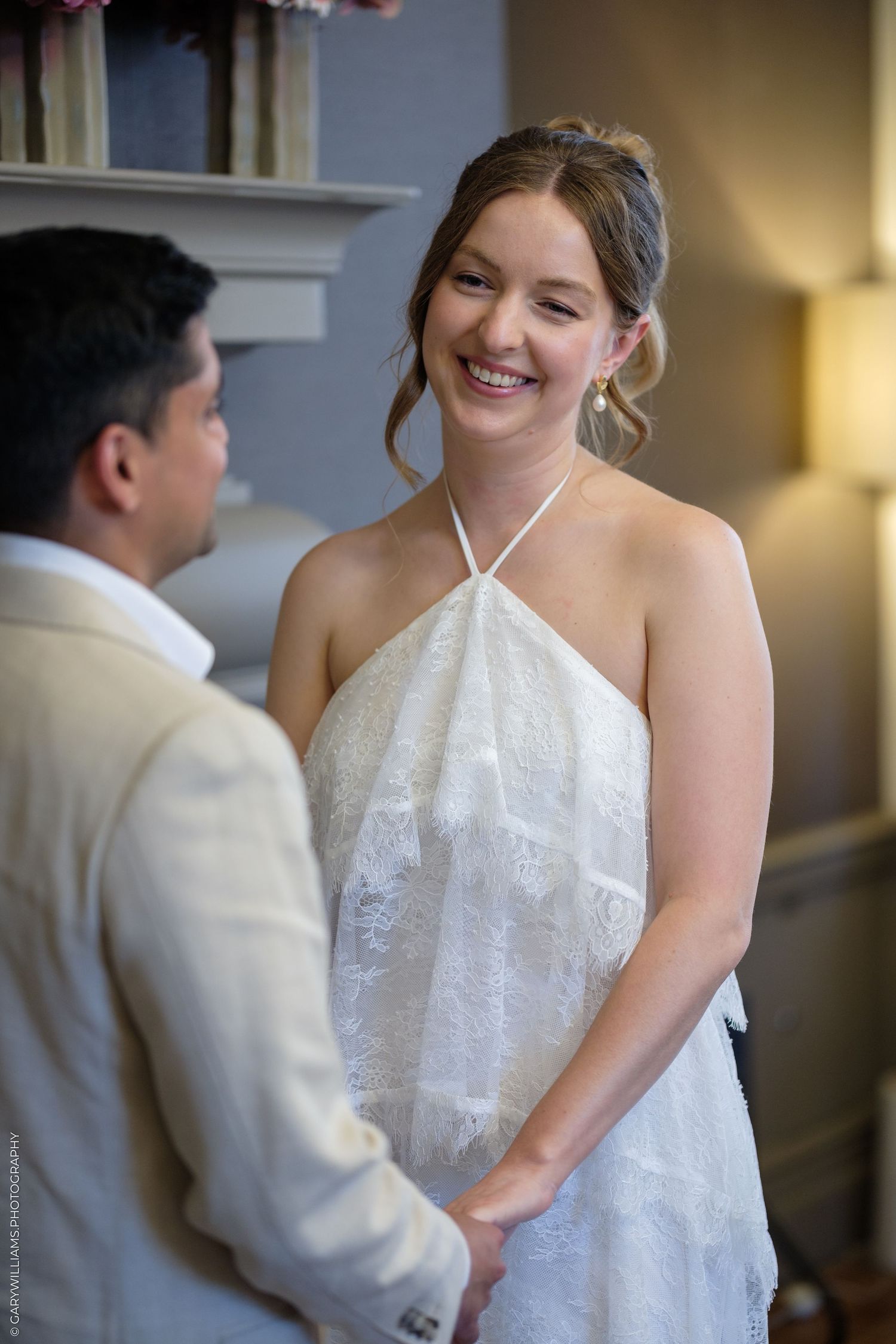 Smiling bride in white halter lace dress holds hands with groom in beige suit during intimate wedding ceremony indoors.