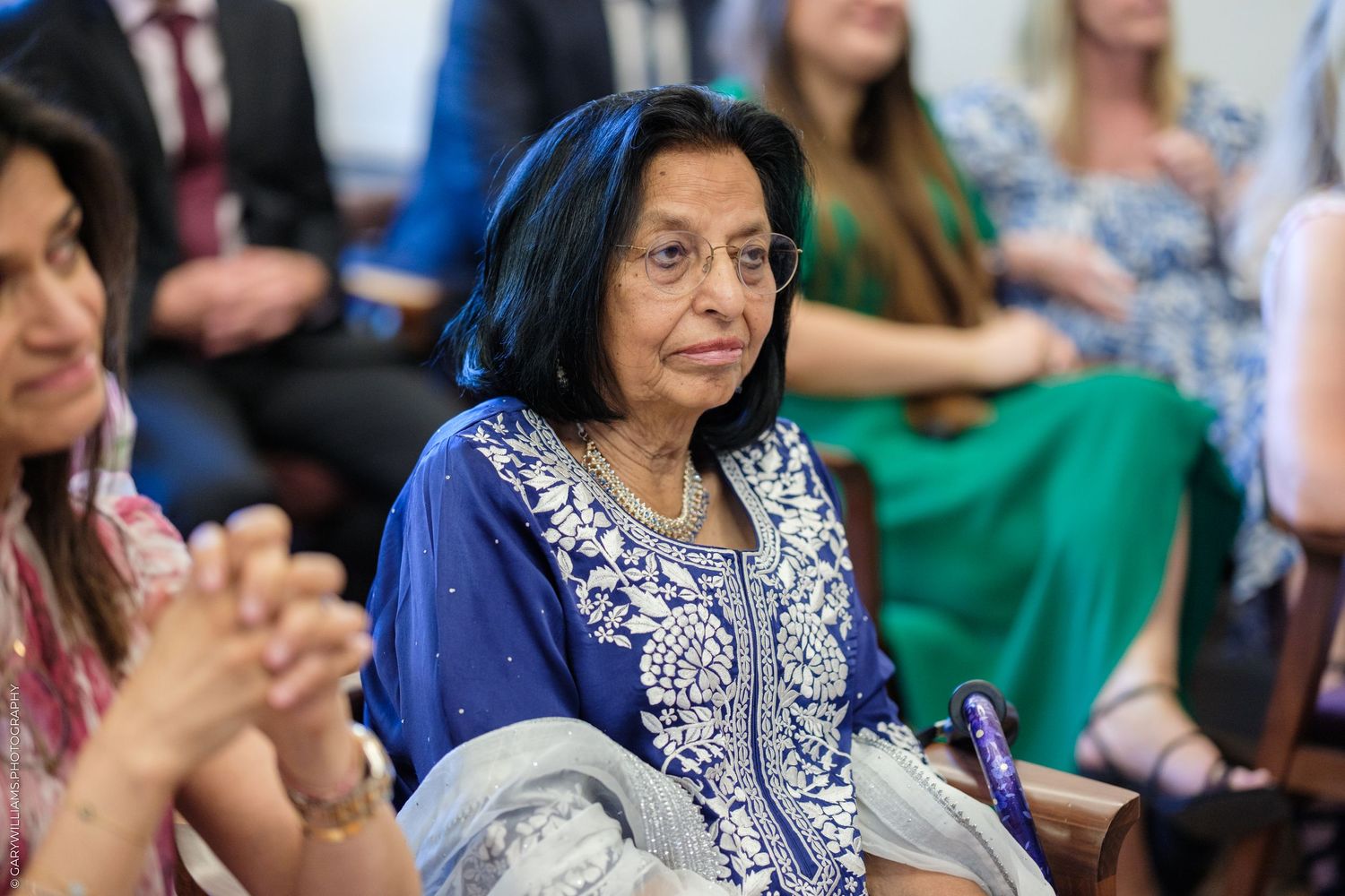 Elderly woman in blue embroidered salwar kameez seated at an indoor gathering, smiling gracefully.
