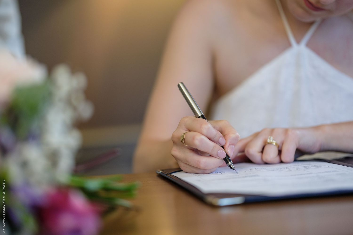 Bride signing wedding marriage certificate document with pen, wearing engagement ring, flowers in foreground.