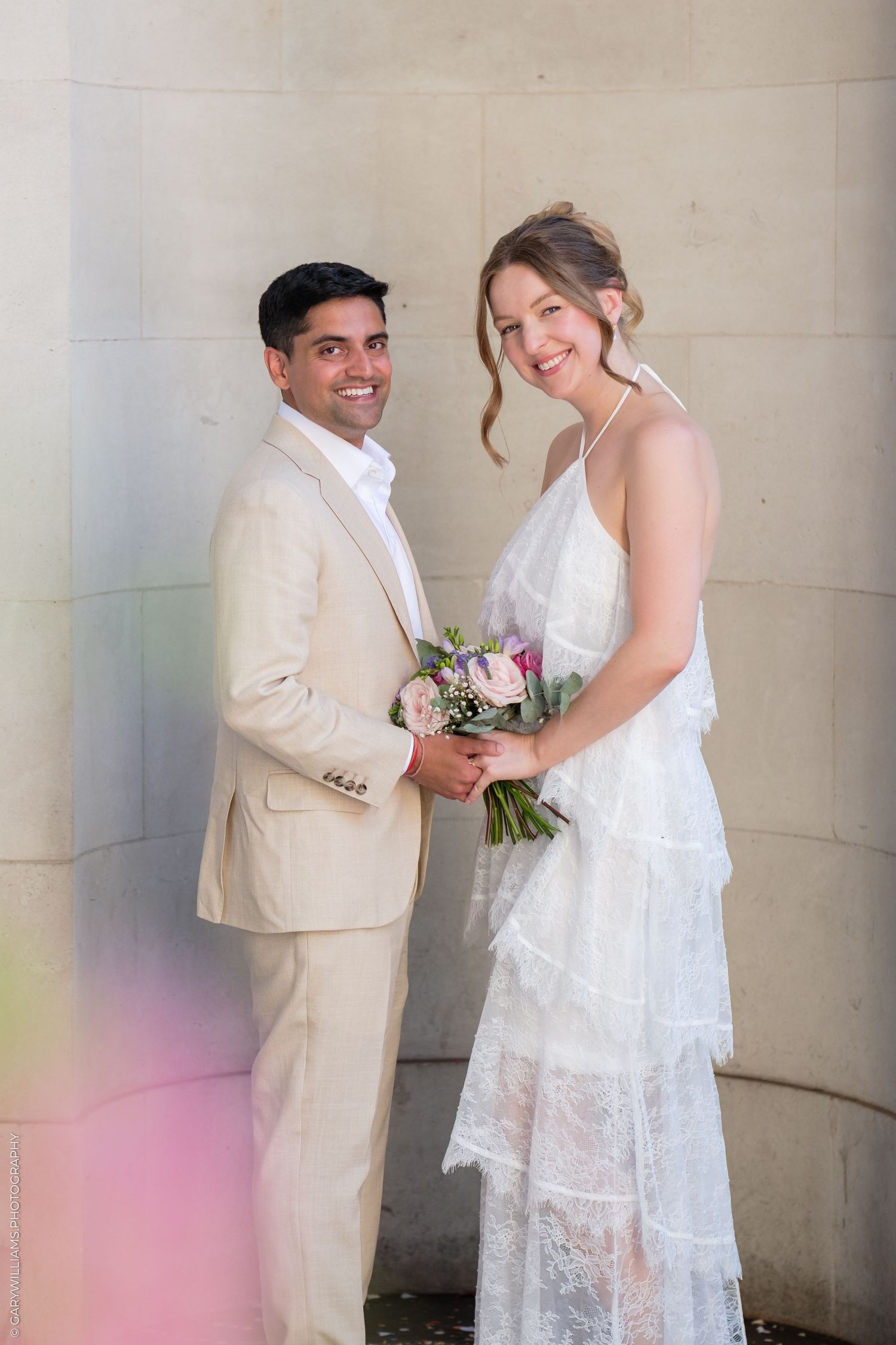 Smiling bride in white lace dress holding bouquet with groom in beige suit posing together at wedding.