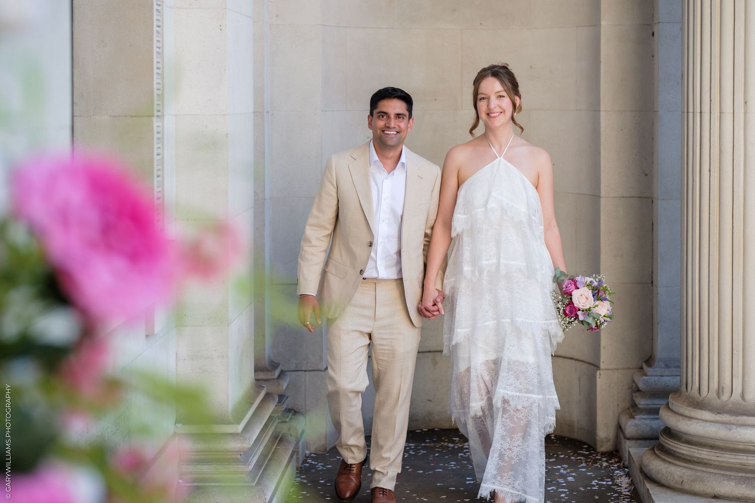 Happy newlywed couple walking hand in hand, bride in white halter dress holding bouquet, groom in beige suit, outdoor venue.