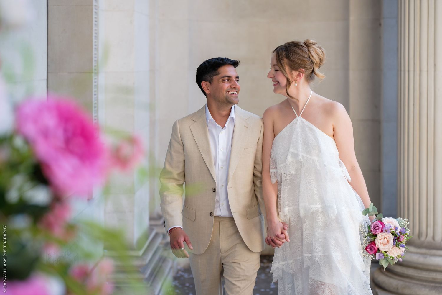 Couple in wedding attire holding hands and smiling among columns, bride holding colorful bouquet.