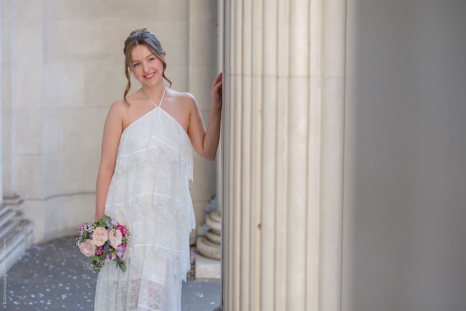 Smiling bride in white halter gown holding bouquet beside stone columns.