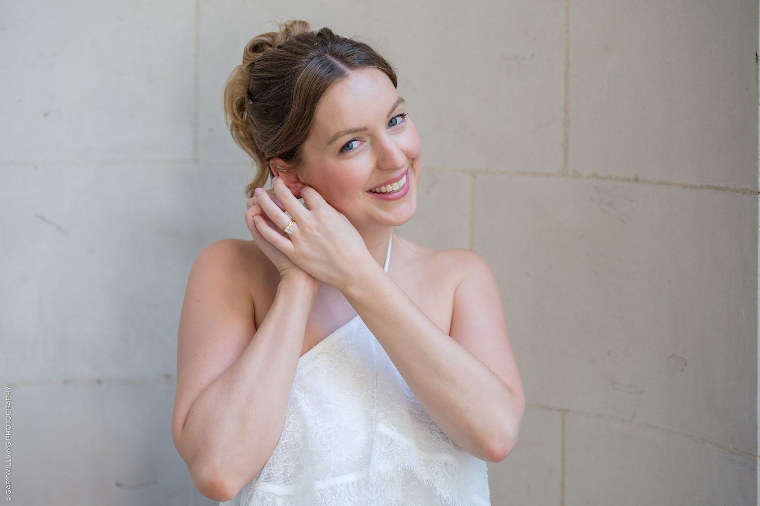 Smiling bride in white dress touching her ear, wearing an engagement ring, posing against a stone wall.