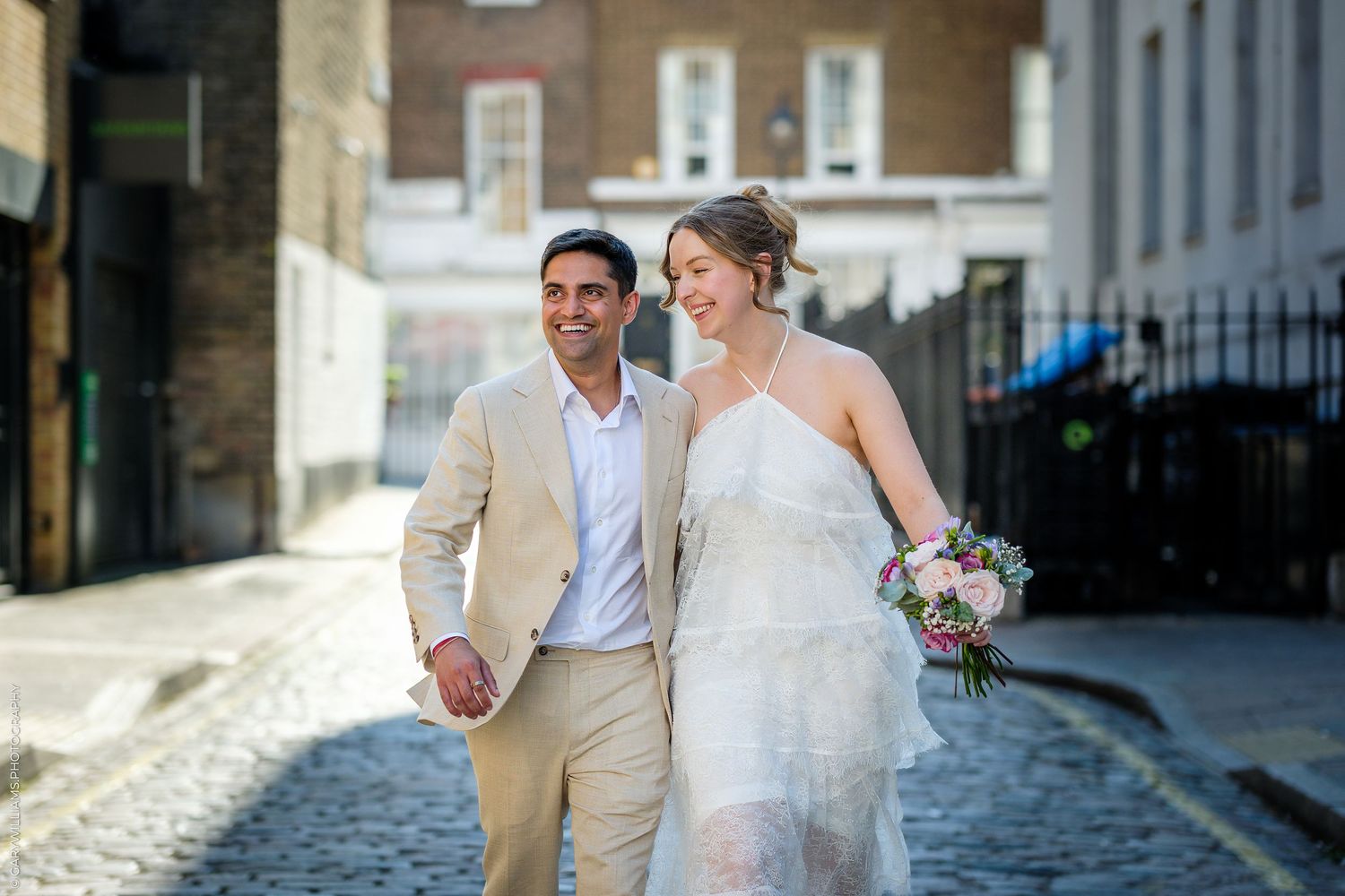 Happy newlyweds walking hand-in-hand on a cobblestone street, bride in white gown with bouquet, groom in beige suit.